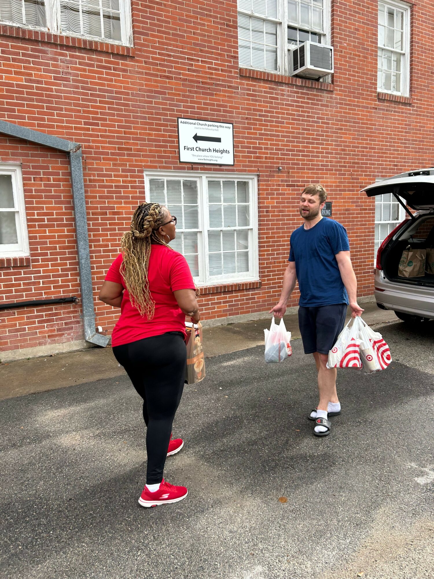 Two women walking on a street in front of a brick building, one in a red shirt and black pants, the other in a blue shirt and shorts, carrying shopping bags.