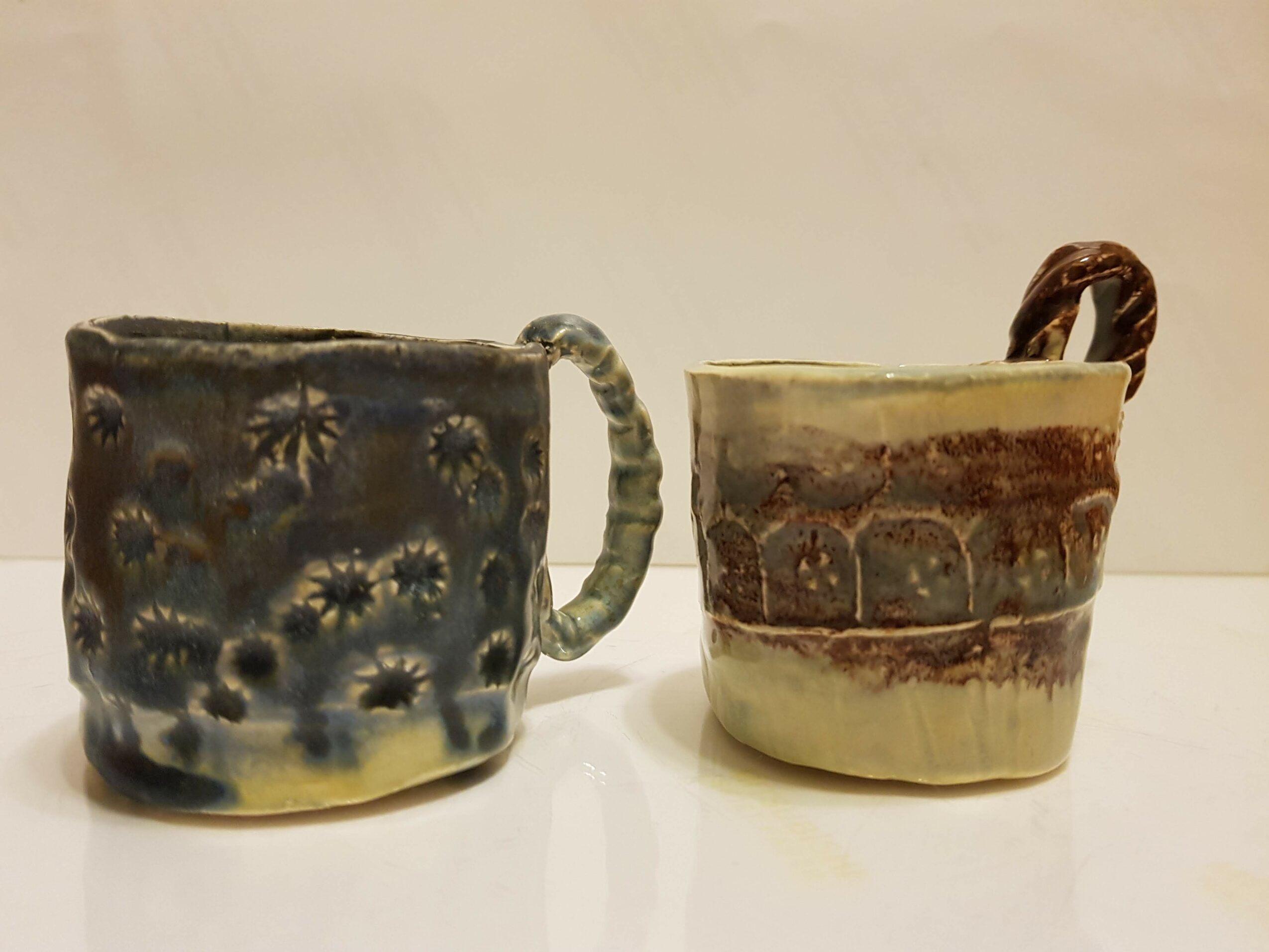 Two ceramic cups with different designs and handles, placed on a white surface against a plain background.