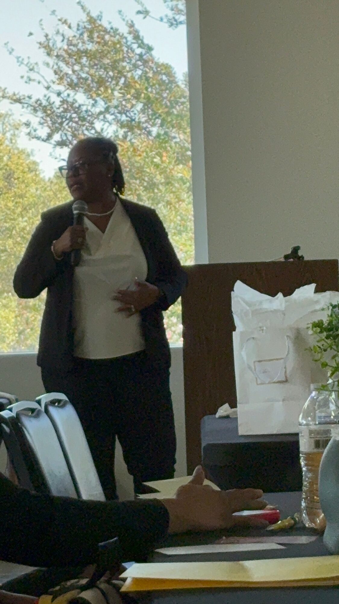 Woman standing and speaking into a microphone in a room with large windows and trees outside.