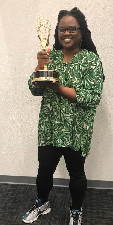 Woman with glasses holding an award trophy, smiling, wearing a green patterned top and black pants, standing against a plain wall.