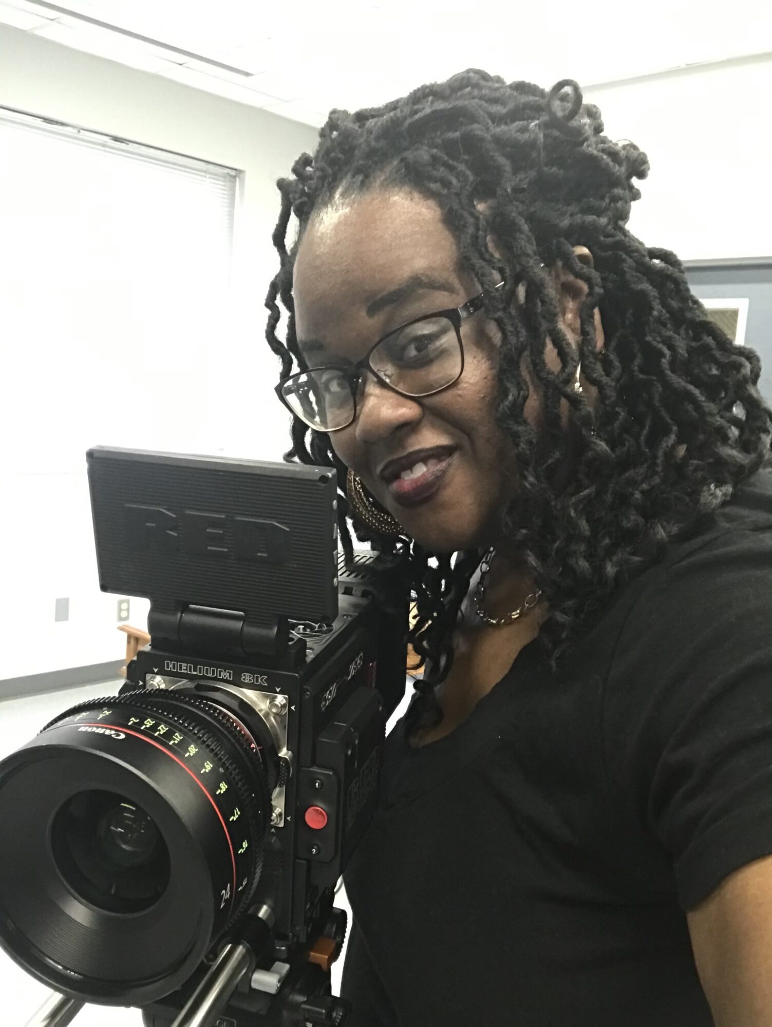 Woman with glasses and dreadlocks holding a camera, smiling, indoors with white walls and a window.