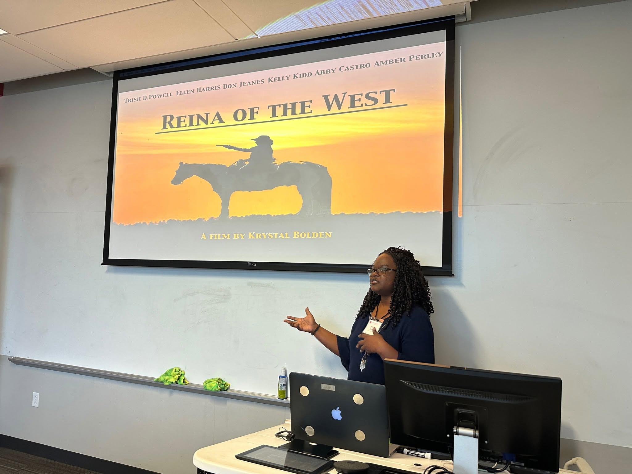 Woman giving presentation in front of a screen displaying a slide titled 'Reina of the West' with a silhouette of a person on a horse at sunset.