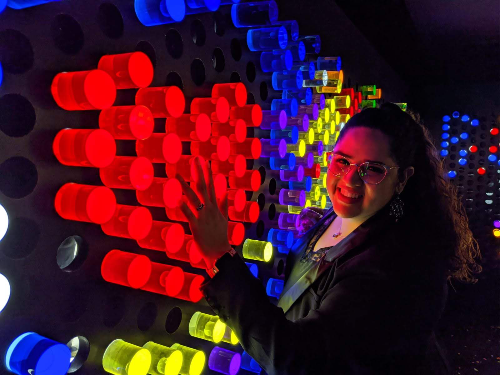 Woman smiling and touching colorful illuminated cylindrical objects on a wall, with vibrant red, yellow, and blue lights.