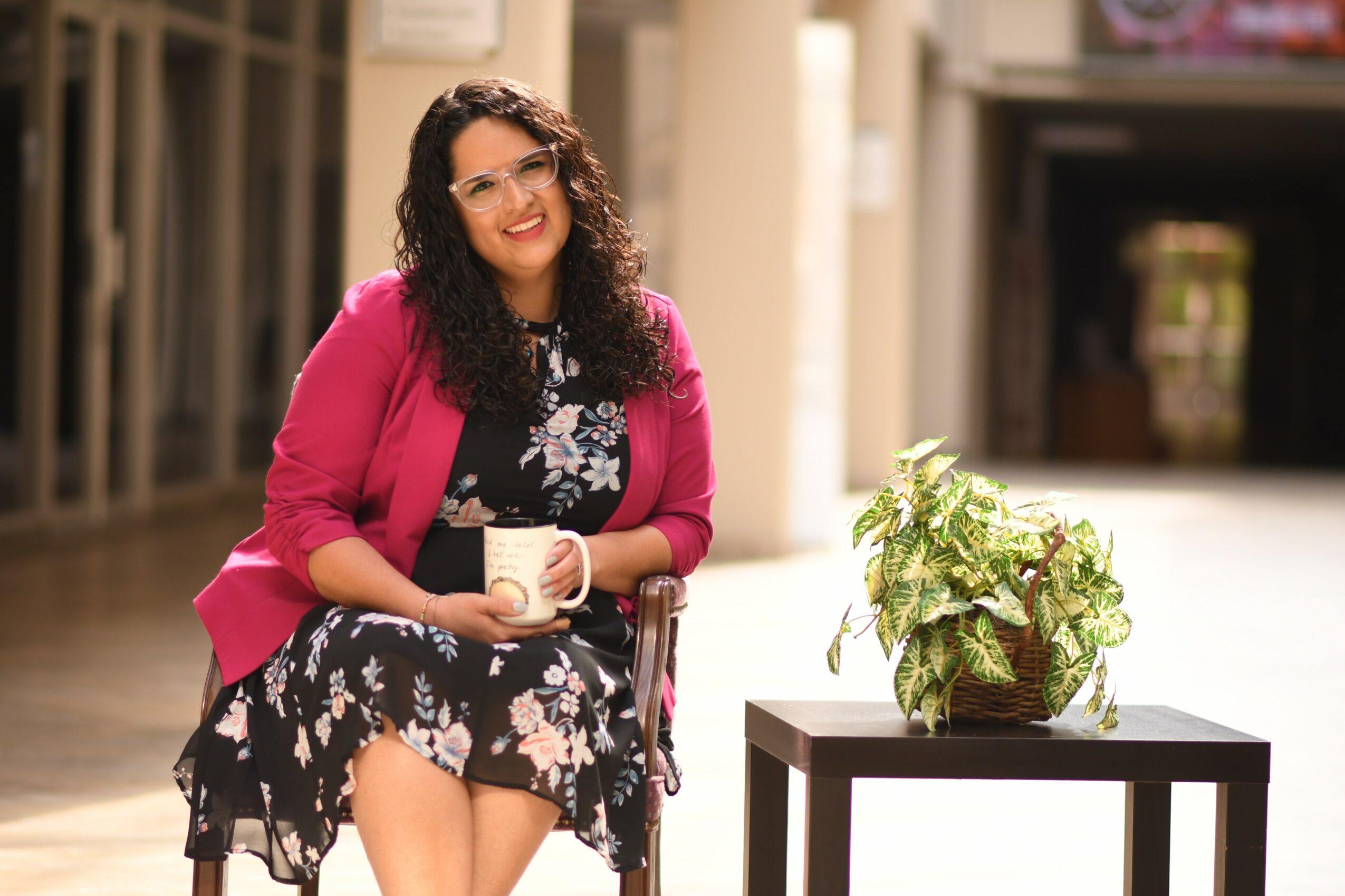 Woman with curly hair in a pink blazer and floral dress holding a mug, standing indoors near a table with a plant.