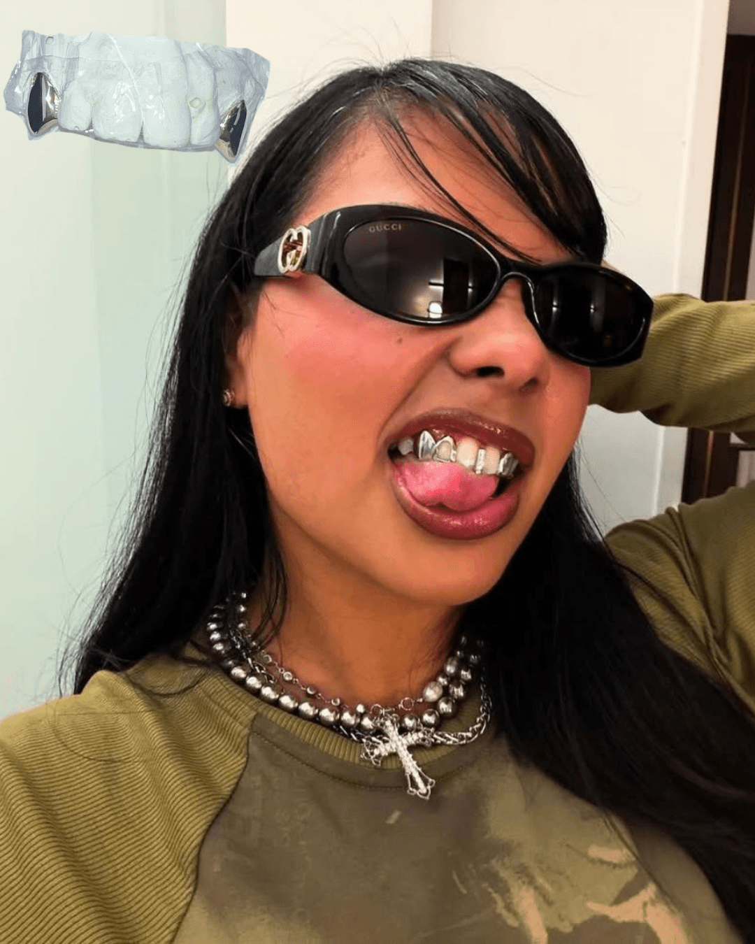 Person with long dark hair wearing sunglasses, sticking out tongue, wearing a green top and layered necklaces.