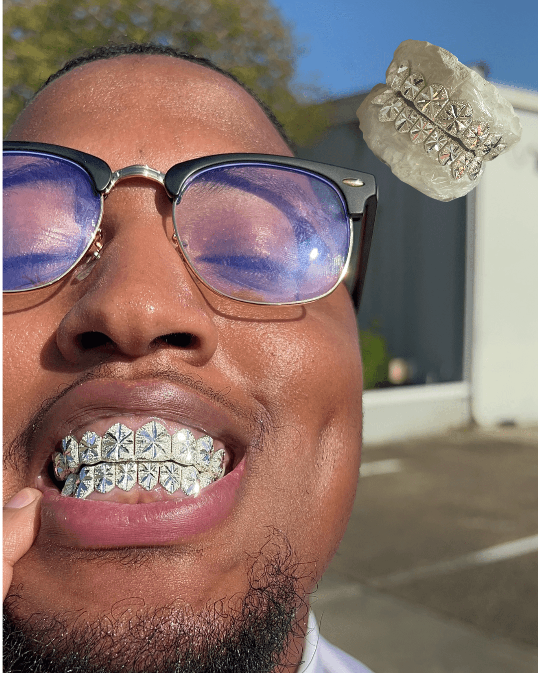 Person with glasses showing teeth with diamond-studded grill, outdoors with blue sky and building in background.