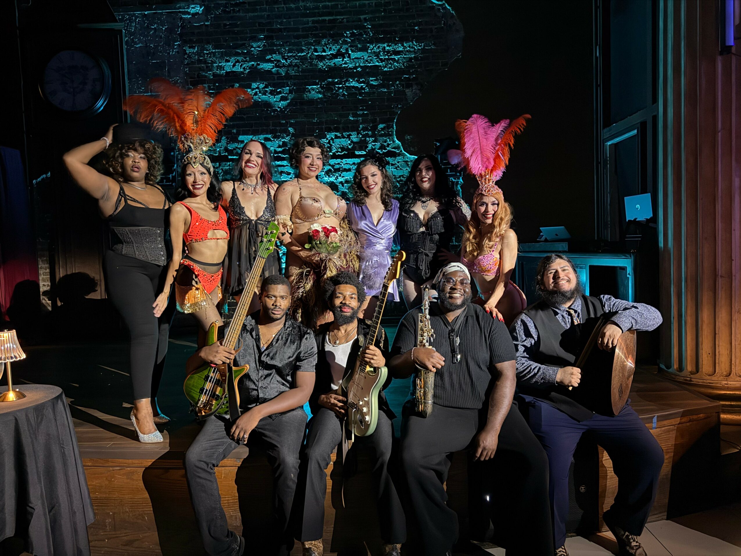 Group of performers on stage, some with feathered headpieces, posing with musical instruments and costumes, in a theater setting.