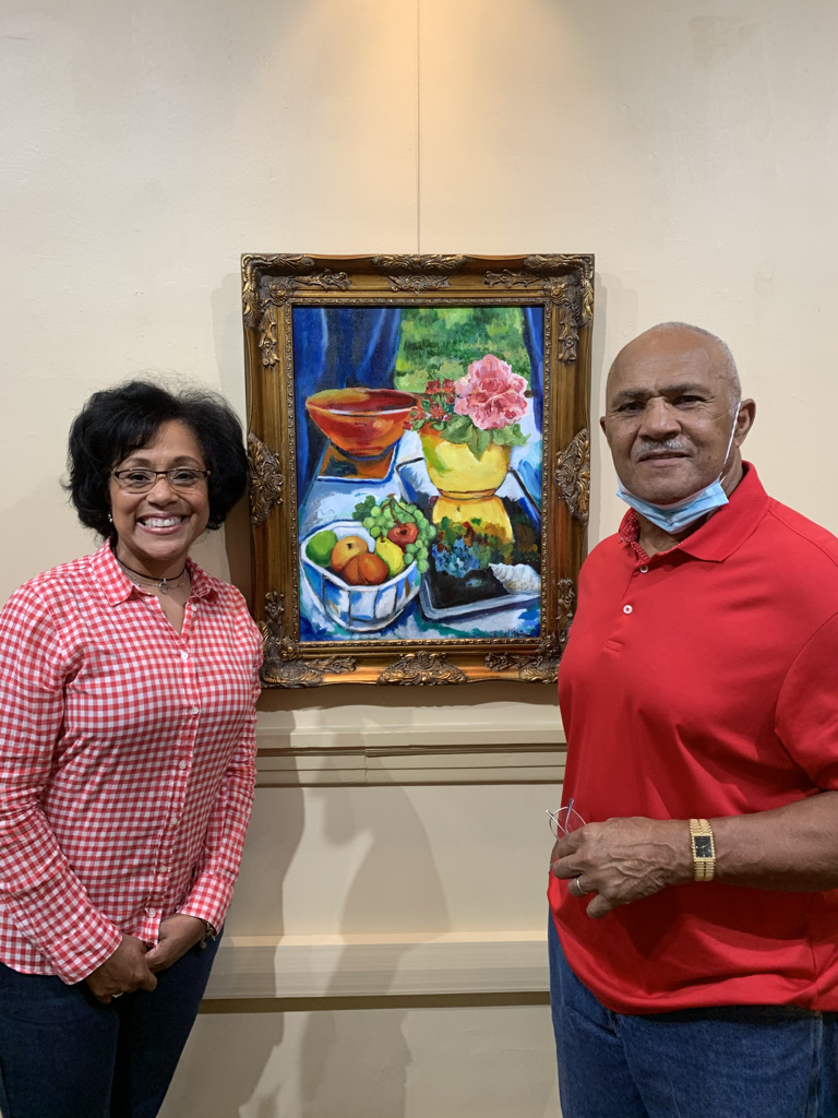 Two people stand in front of a colorful still life painting on a wall, smiling at the camera.