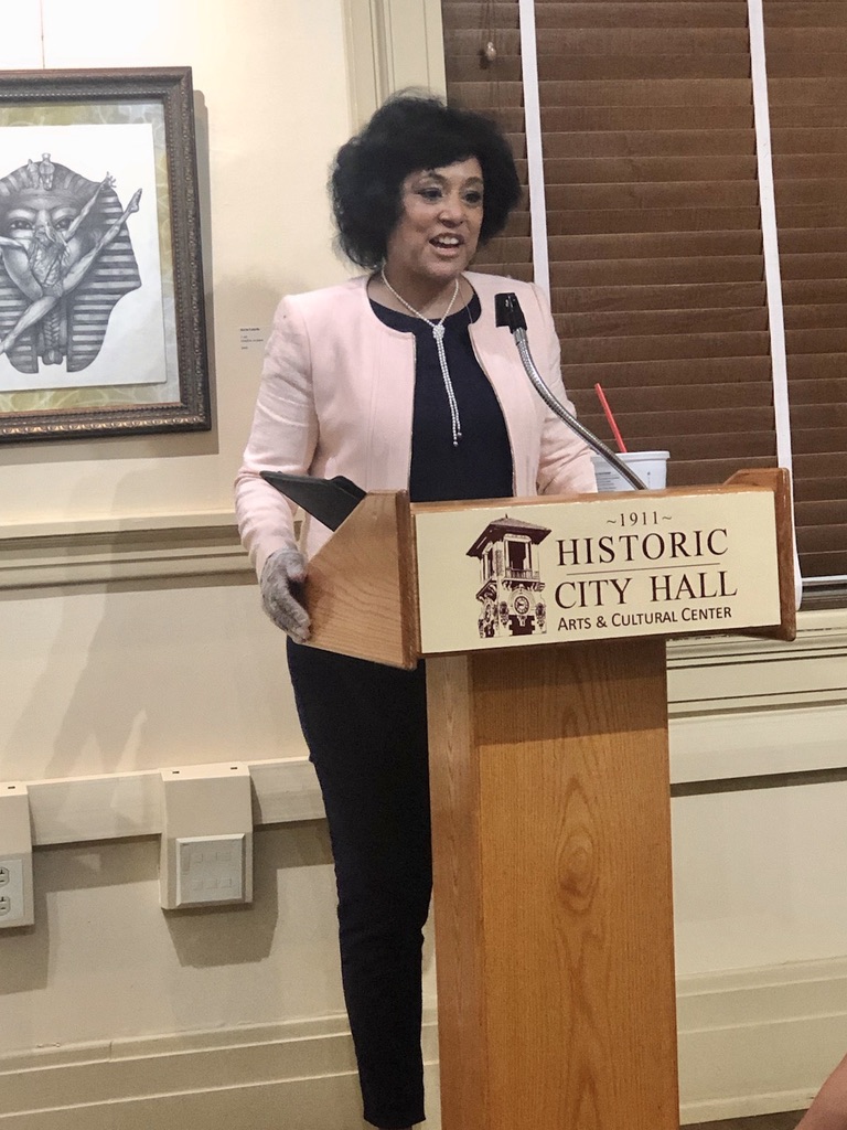 Woman speaking at a podium in a room with framed artwork and wooden blinds, wearing a light pink blazer and dark pants.