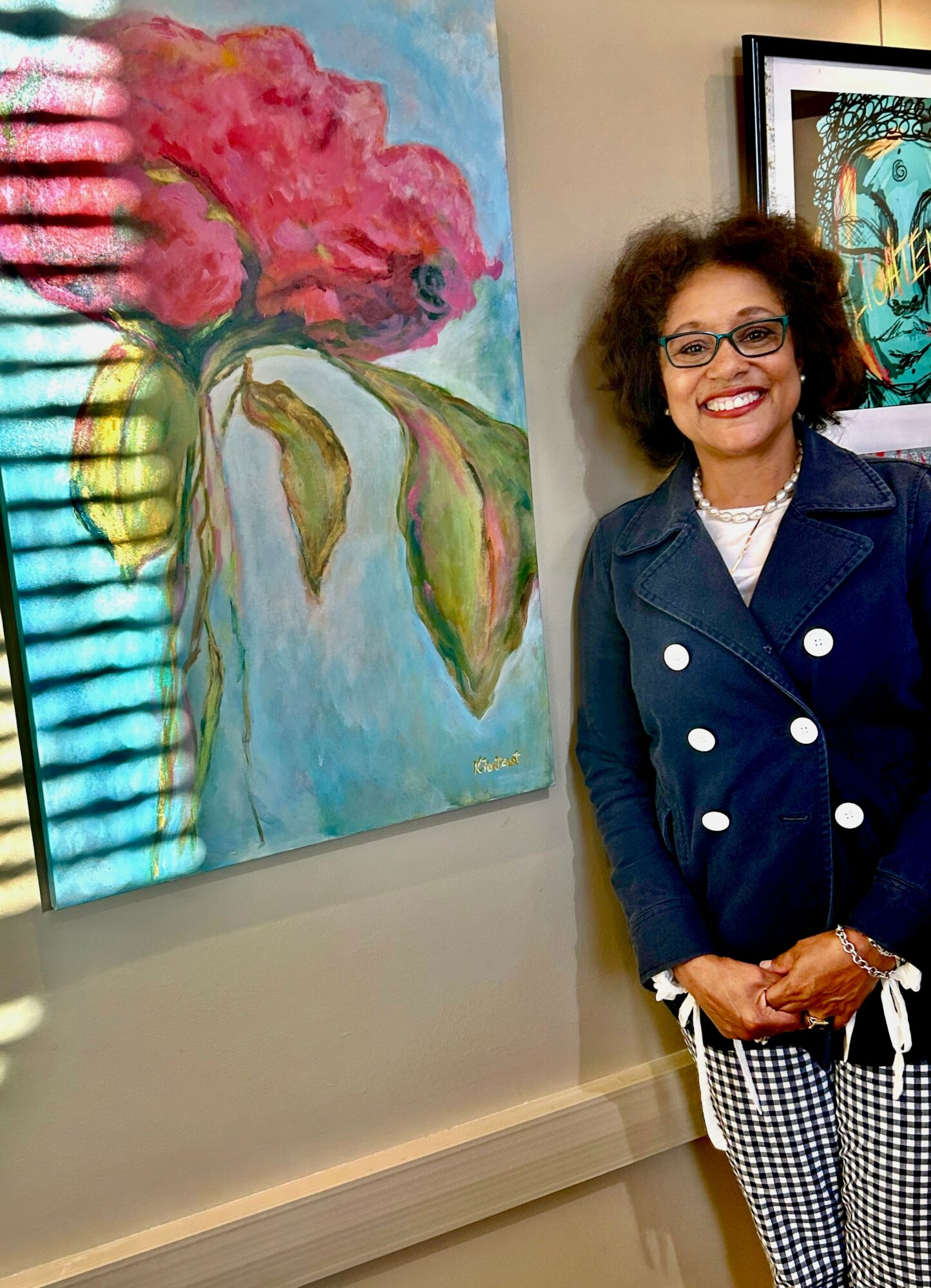 Smiling woman with curly hair and glasses stands next to colorful floral artwork in gallery.