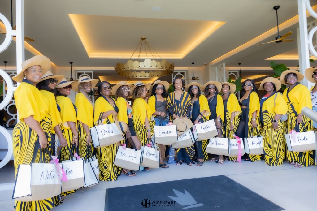 Group of women in yellow and black outfits with hats, holding shopping bags, standing indoors with a chandelier and ceiling lights.