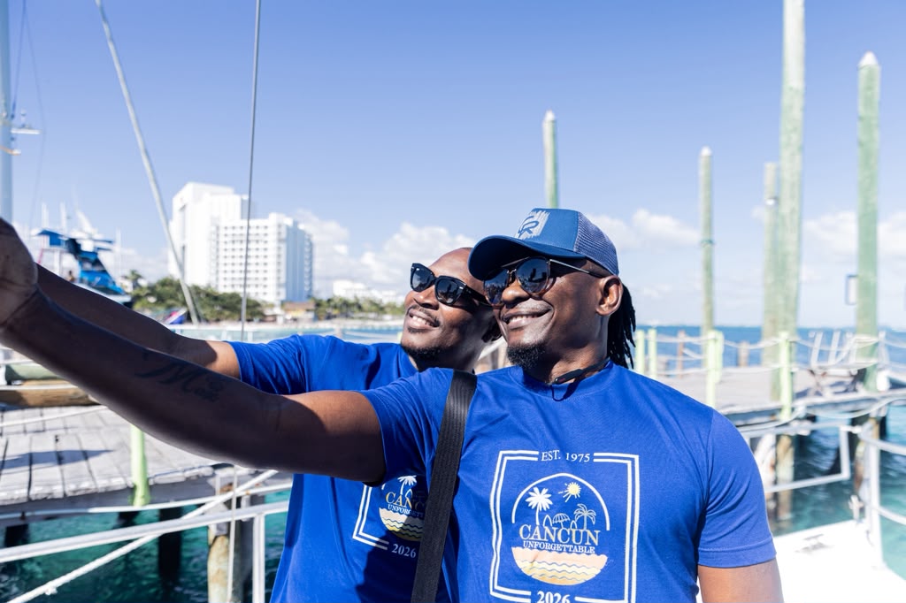 Two people in blue shirts and sunglasses taking a selfie at a marina with boats and tall buildings in the background.