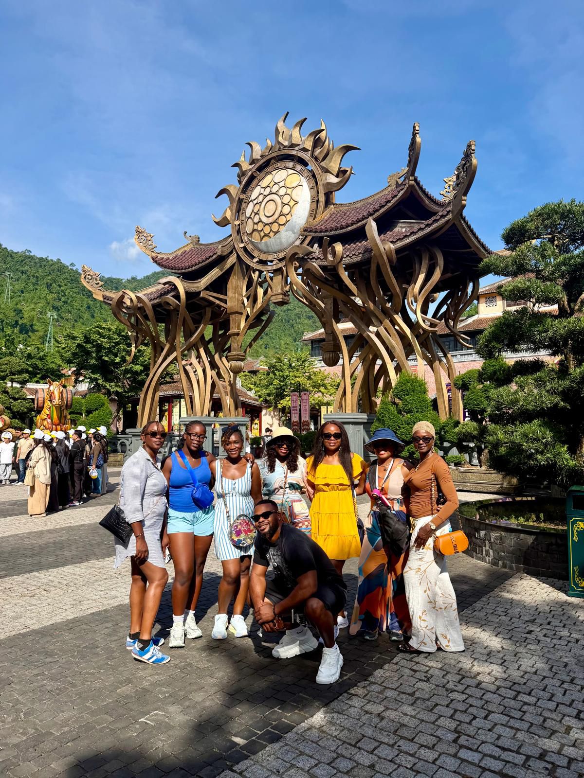 Group of seven people standing in front of an ornate, large wooden structure with a circular design and decorative elements, outdoors on a sunny day.