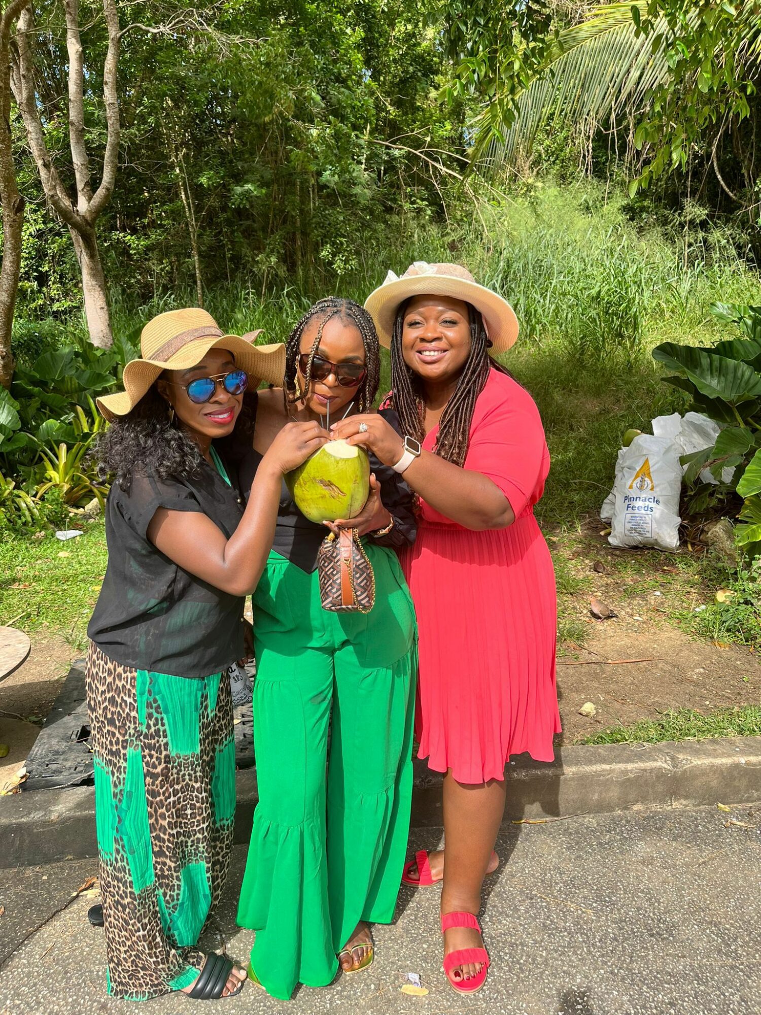 Three women standing outdoors in a lush green area, smiling and holding a yellow coconut, wearing hats and colorful clothing.