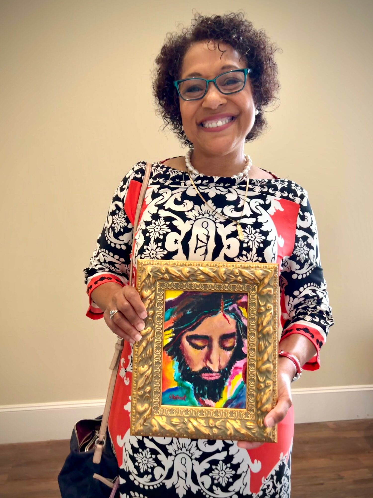 Smiling woman with glasses holding a framed colorful painting of a face, standing indoors against a plain wall.
