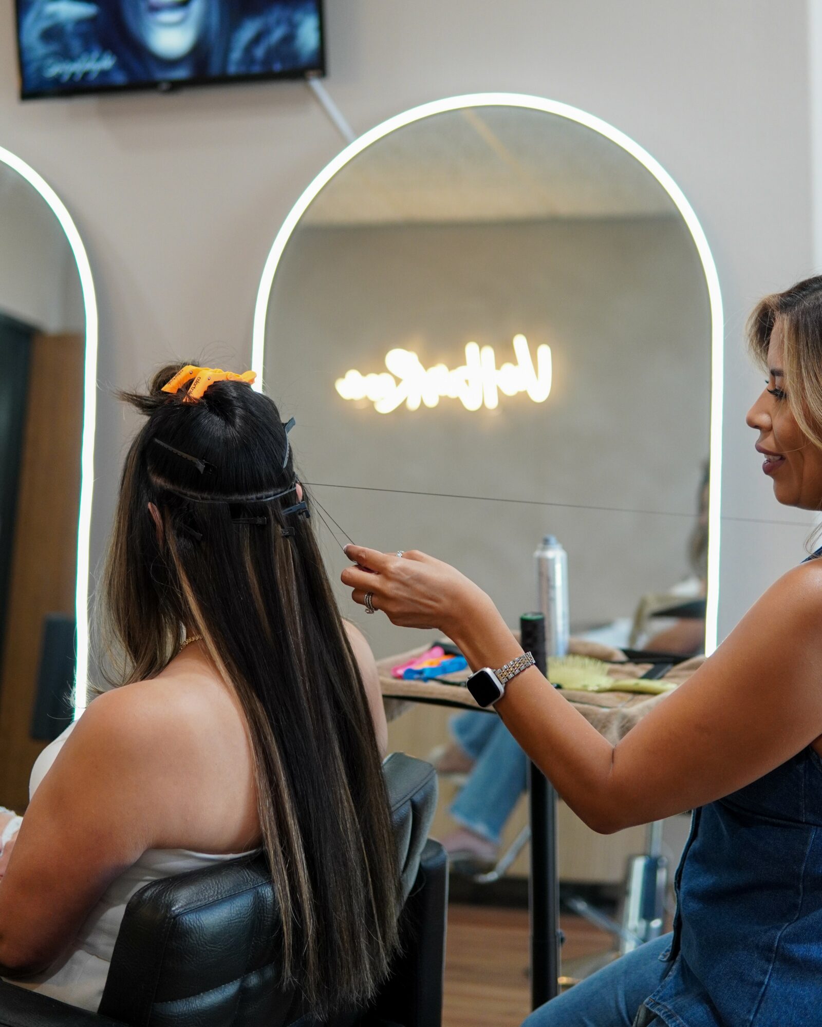 A woman with long dark hair is getting her hair styled in a salon, with mirrors and salon tools visible.