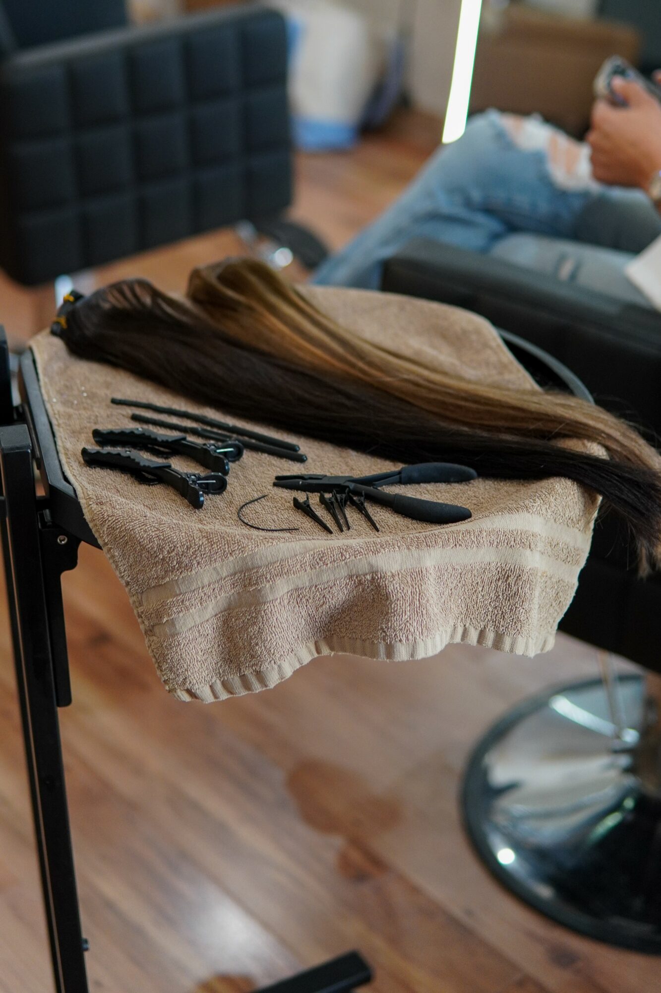 Long hair and hair styling tools on a towel, with a person sitting nearby in a salon setting.