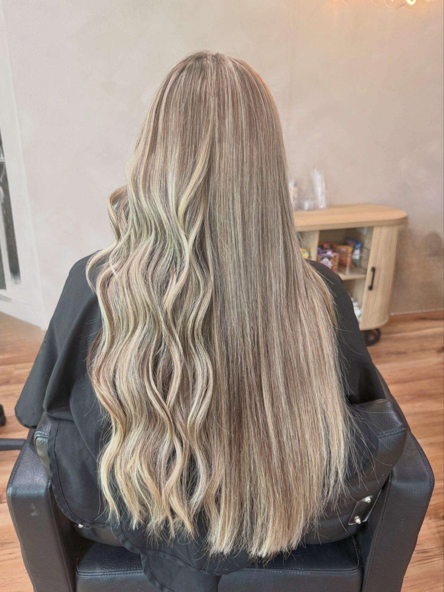 Back view of a person with long, wavy, blonde hair sitting in a salon chair, with a wooden table in the background.
