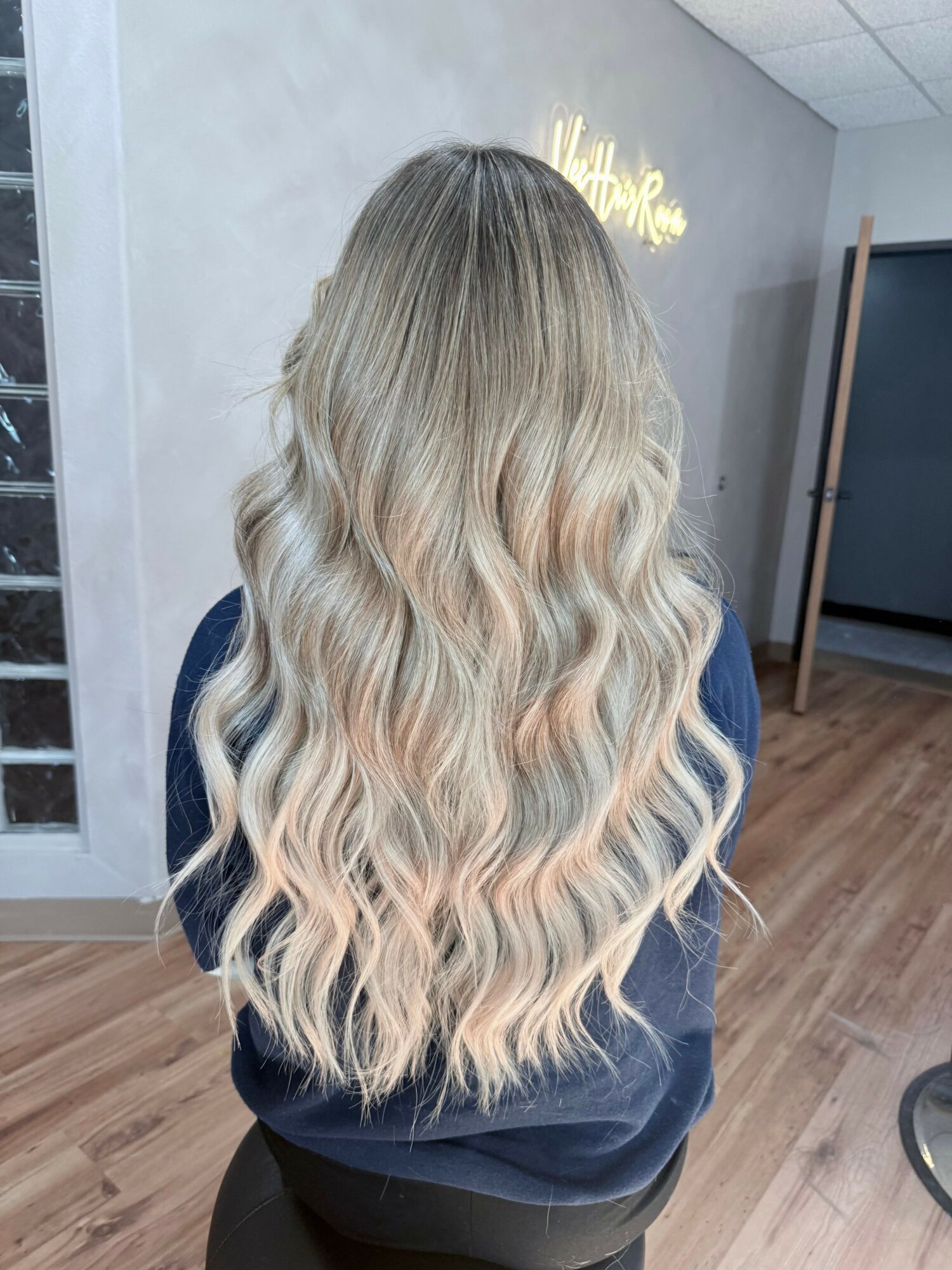 Back view of a woman with long, wavy, blonde hair in a salon, sitting on a stool, with a neon sign in the background.