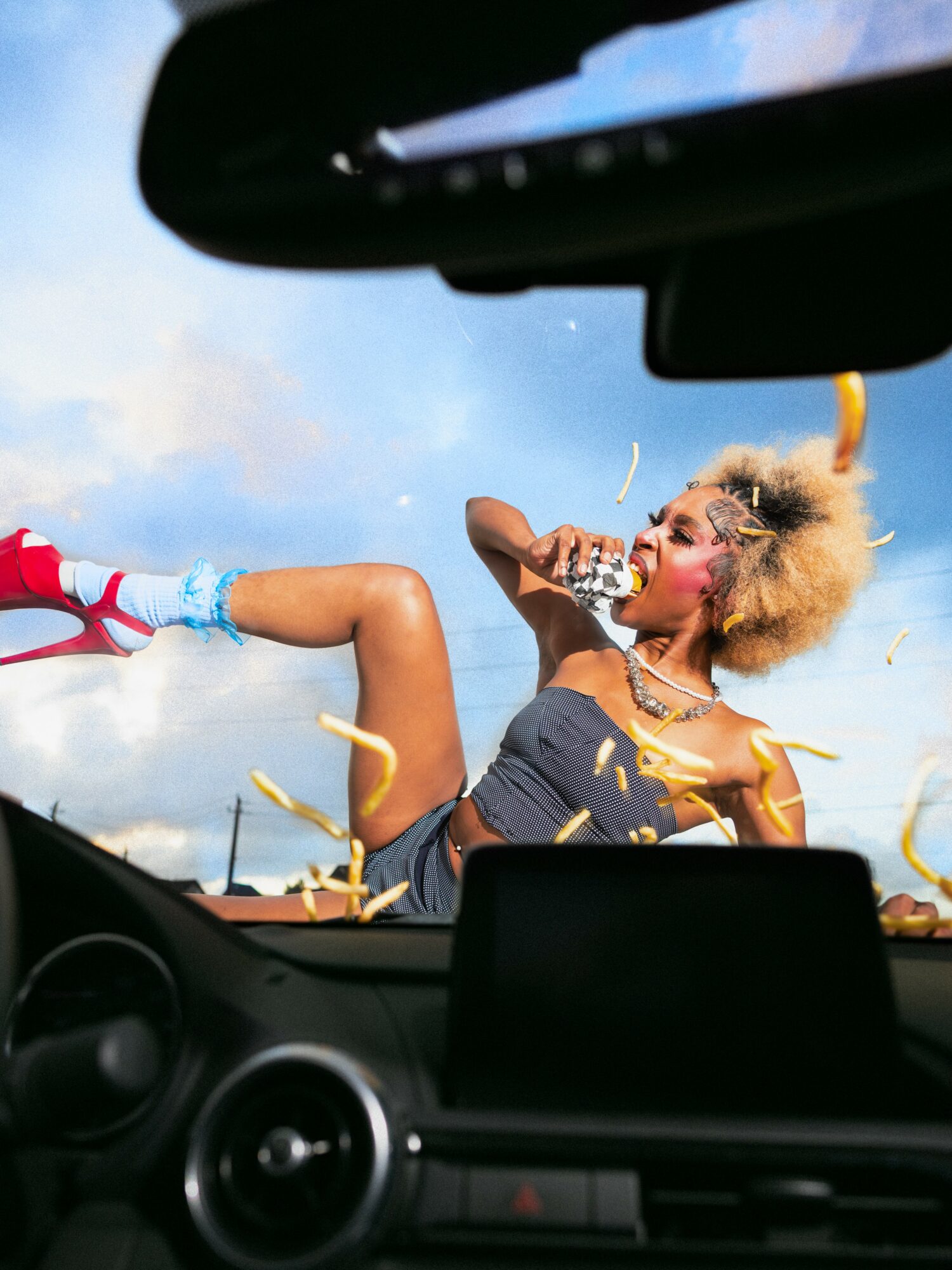 Person with curly hair lying on a car dashboard, surrounded by confetti, with a blue sky background.