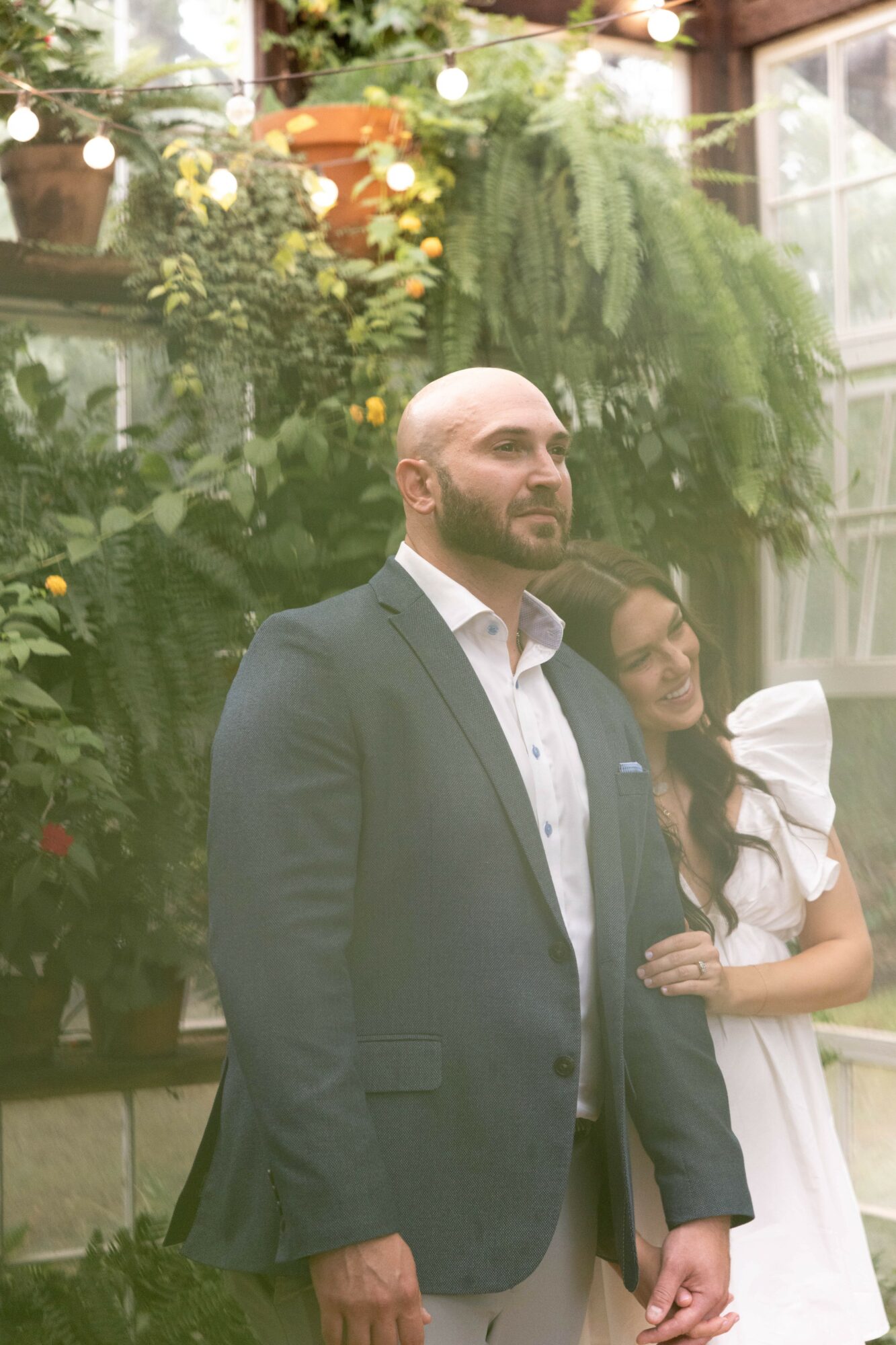 A man with a beard in a suit stands in front of greenery, with a woman in a white dress beside him, smiling.
