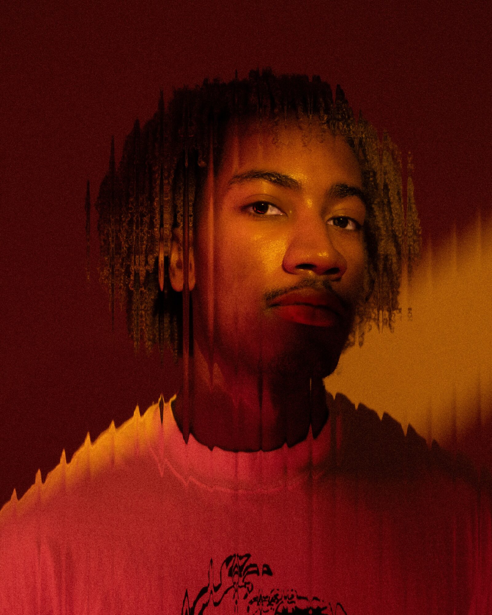 Portrait of a young person with dreadlocks, looking at the camera, with warm lighting and a dark background.