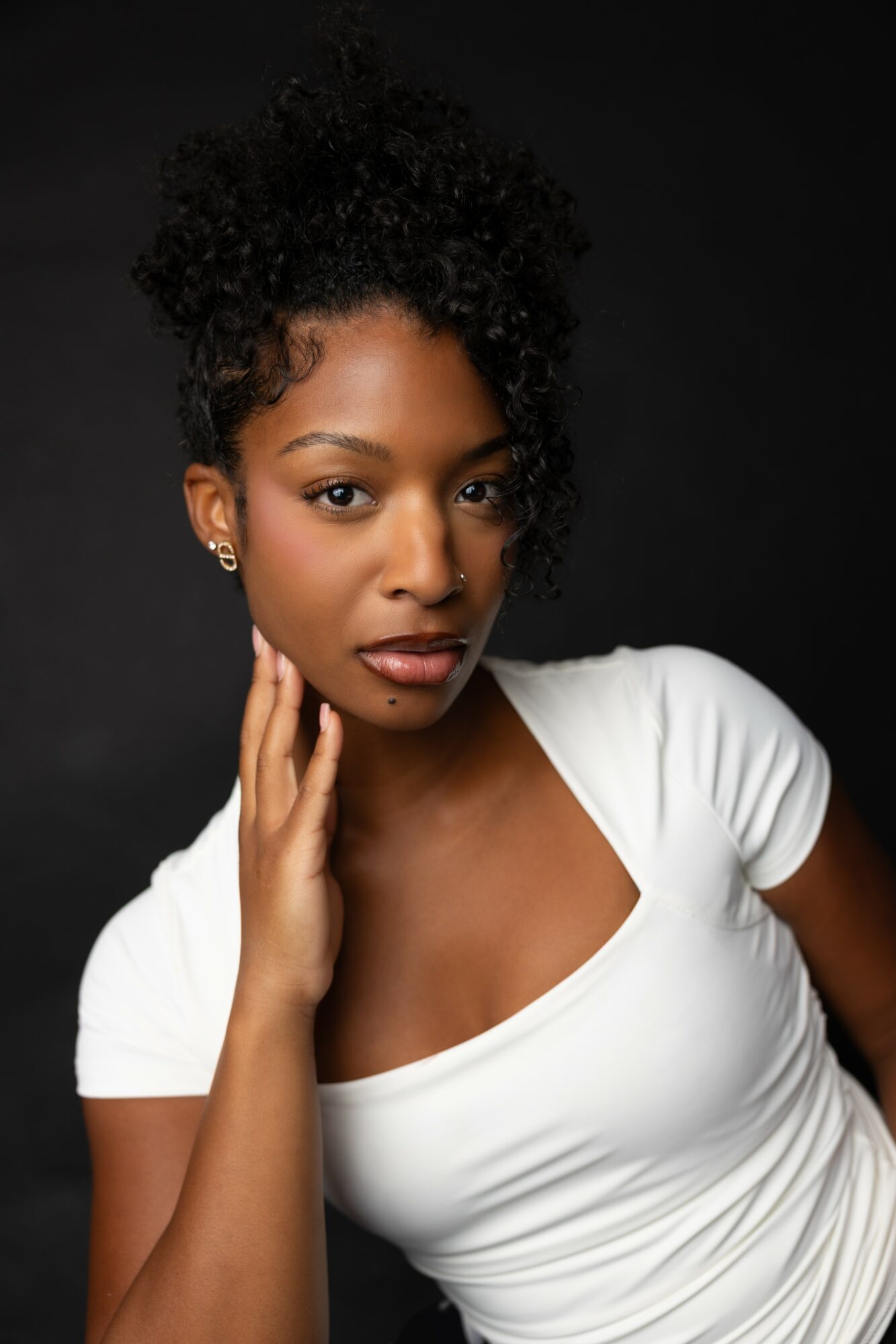 Portrait of a woman with natural curly hair, wearing a white top, touching her face, against a dark background.