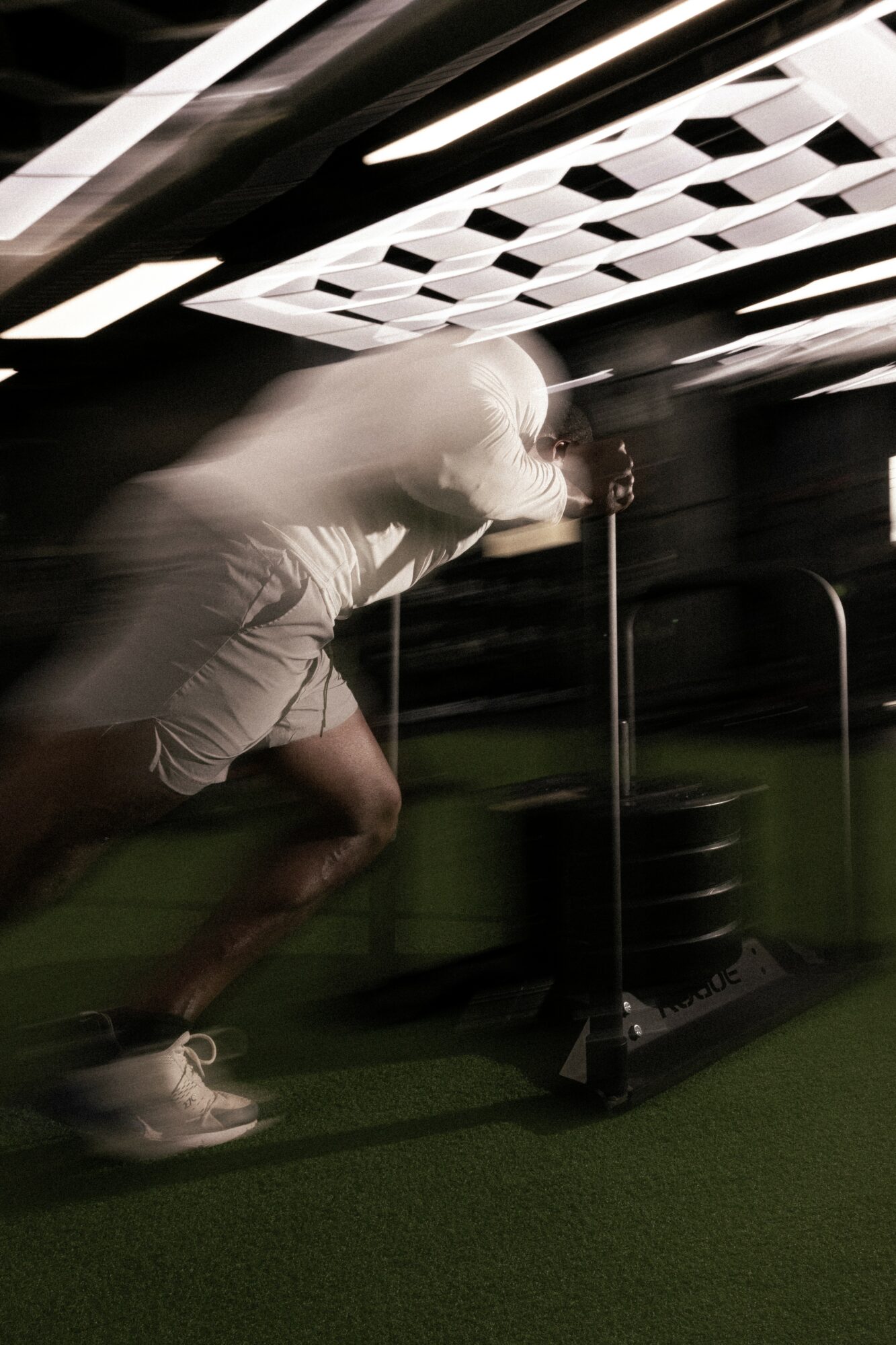 Person pushing a sled indoors with a ceiling featuring a geometric pattern, blurred motion, and green flooring.