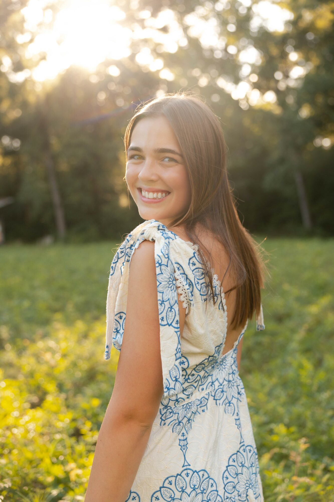 Smiling woman with long hair outdoors in sunlight, wearing a patterned dress, standing in a grassy area with trees.