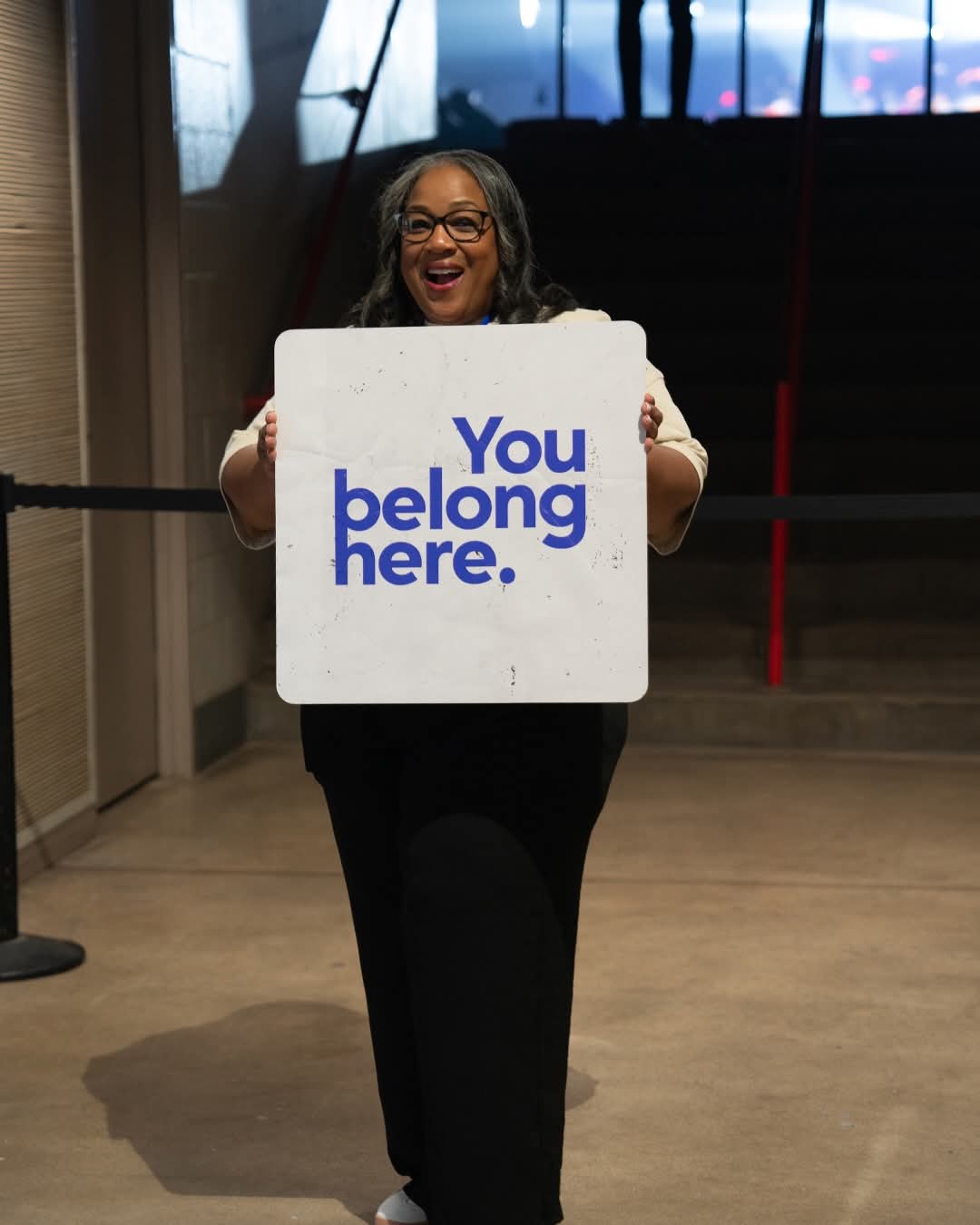 Smiling woman holding a sign that says 'You belong here' in a modern indoor space.