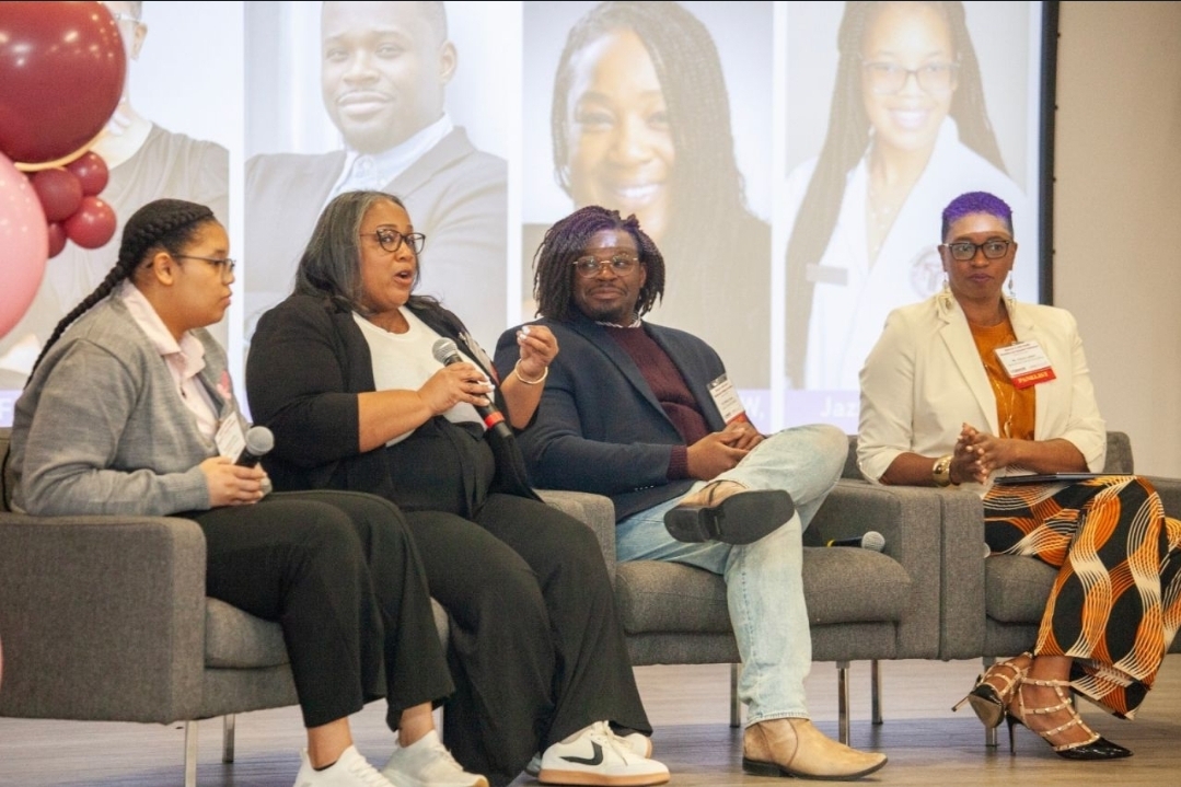 Four women sitting on a panel, one speaking into a microphone, with a large screen showing faces behind them.