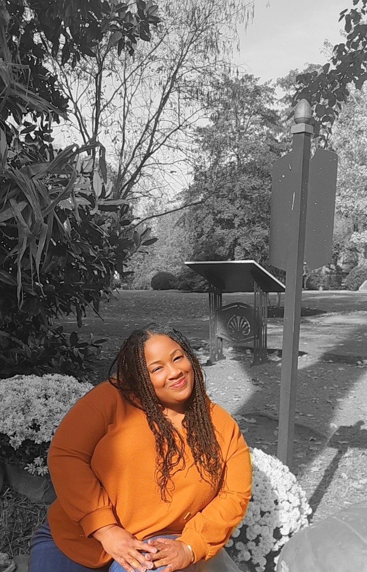 Smiling woman with long curly hair wearing an orange top outdoors, trees and a bench in background.