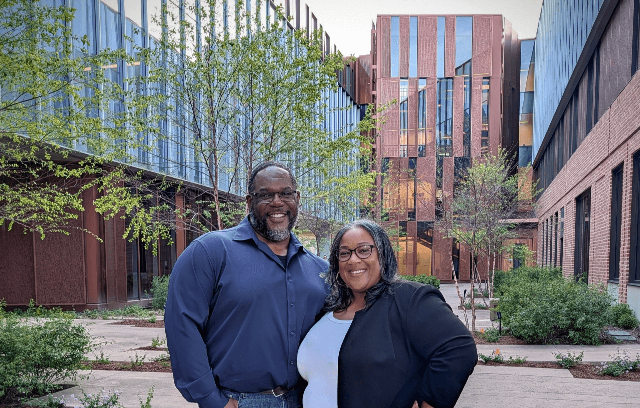 Two people standing outdoors in an urban area with modern buildings and trees, smiling at the camera.