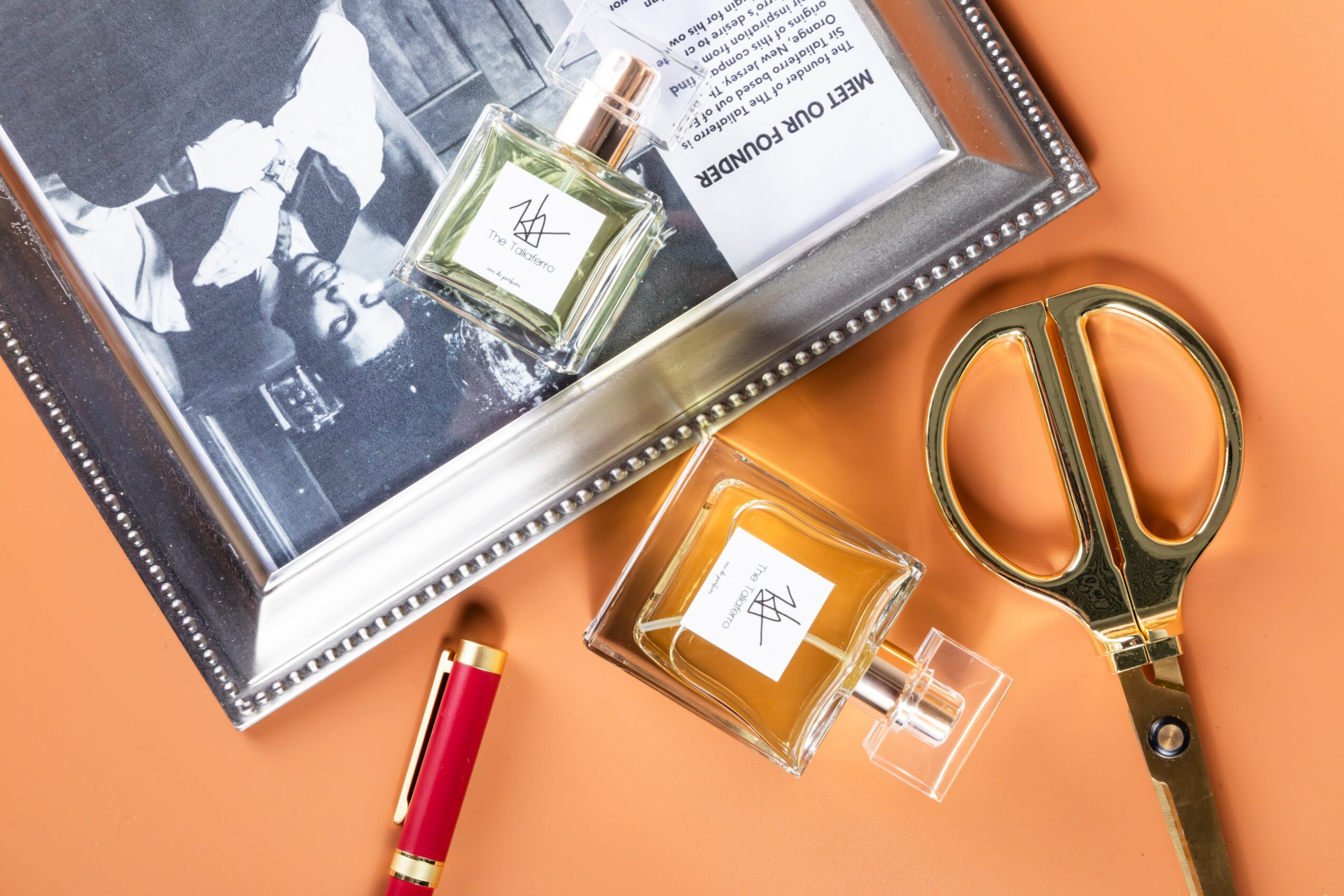 A glass perfume bottle with a gold cap, a pair of gold scissors, and a framed black-and-white photo on an orange surface.