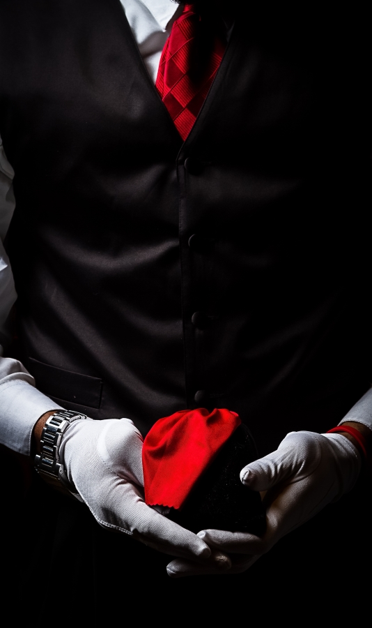 Person in black suit holding a red heart-shaped object with white gloves, dark background.