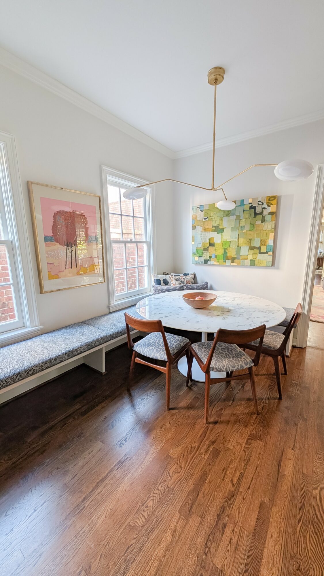 Dining room with round table, four chairs, artwork on walls, window seat, and hardwood floor.