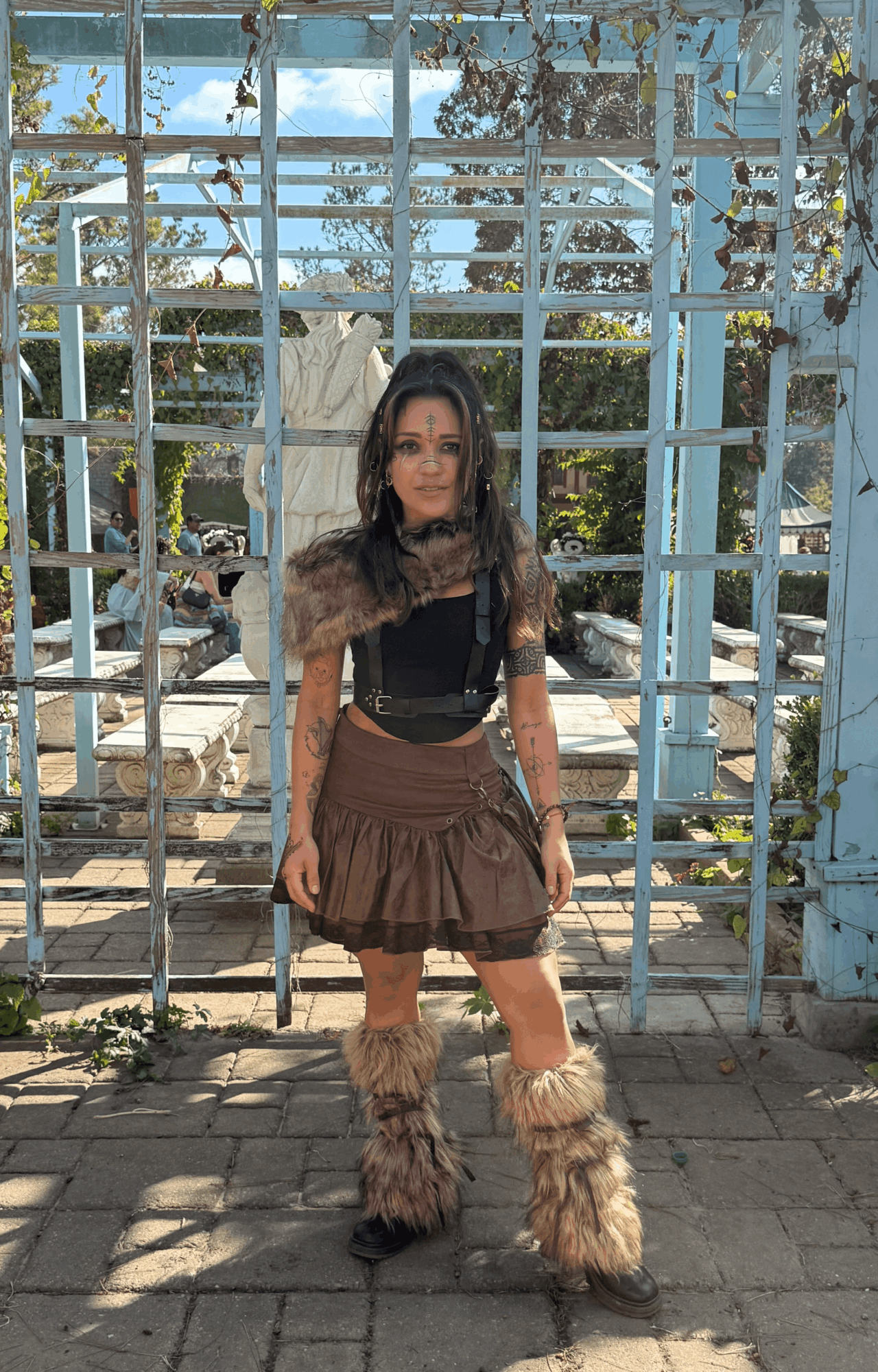 Woman standing outdoors in front of a decorative gate, wearing furry leg warmers and a fur stole, with trees and sky in background.
