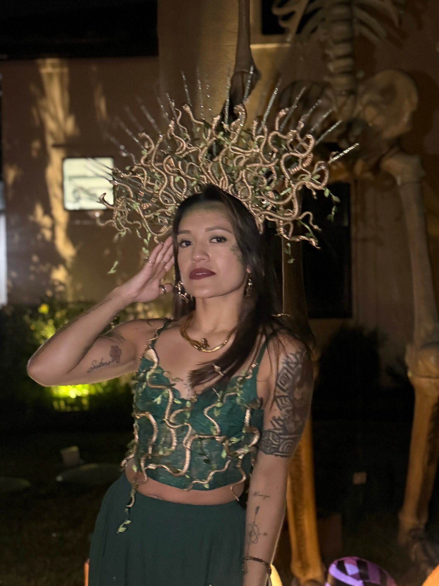 Woman wearing an ornate headpiece and a green outfit, standing indoors with dim lighting and decorative elements.