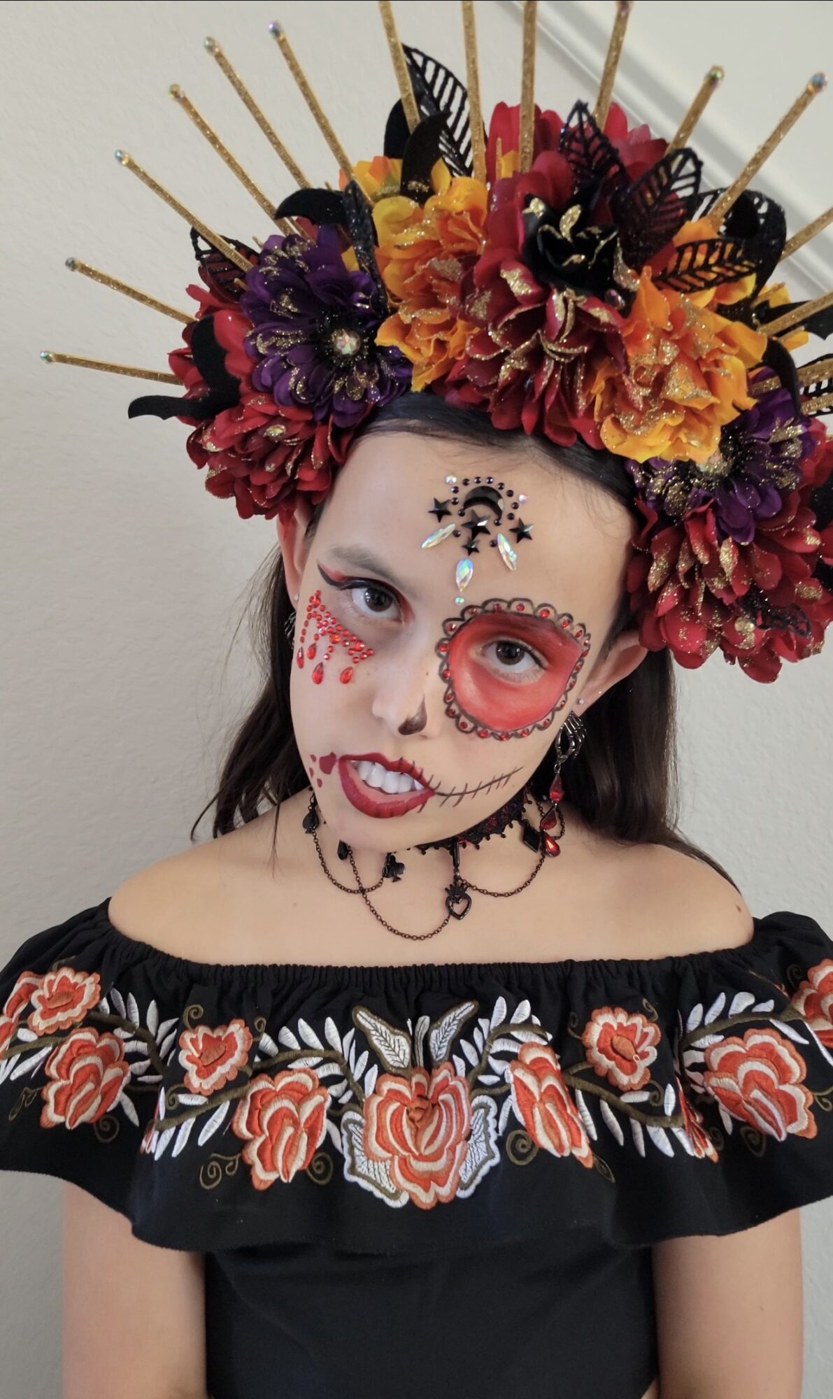 Person with face painted in sugar skull style, wearing a floral off-shoulder top and a large floral headdress with sticks.