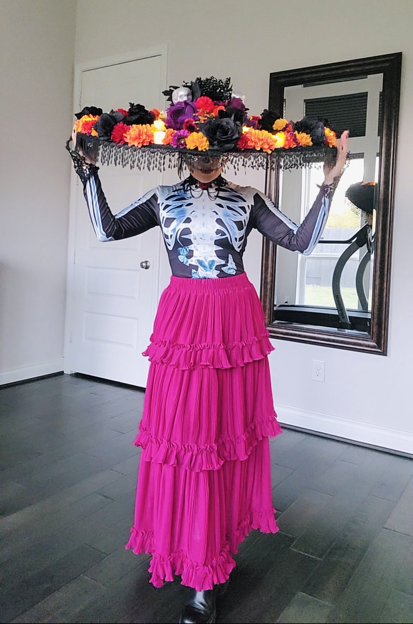 Person holding a large floral hat, wearing a patterned top and bright pink layered skirt, standing indoors near a mirror.
