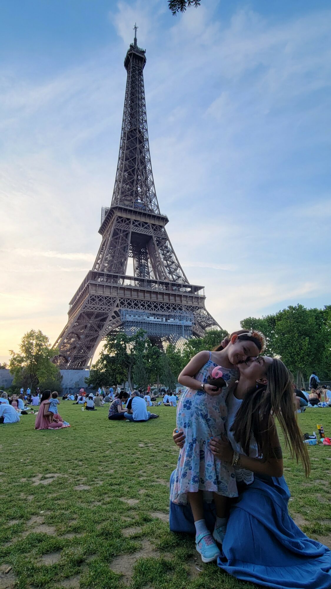 Two people embrace in front of the Eiffel Tower with a crowd sitting on grass, blue sky overhead.