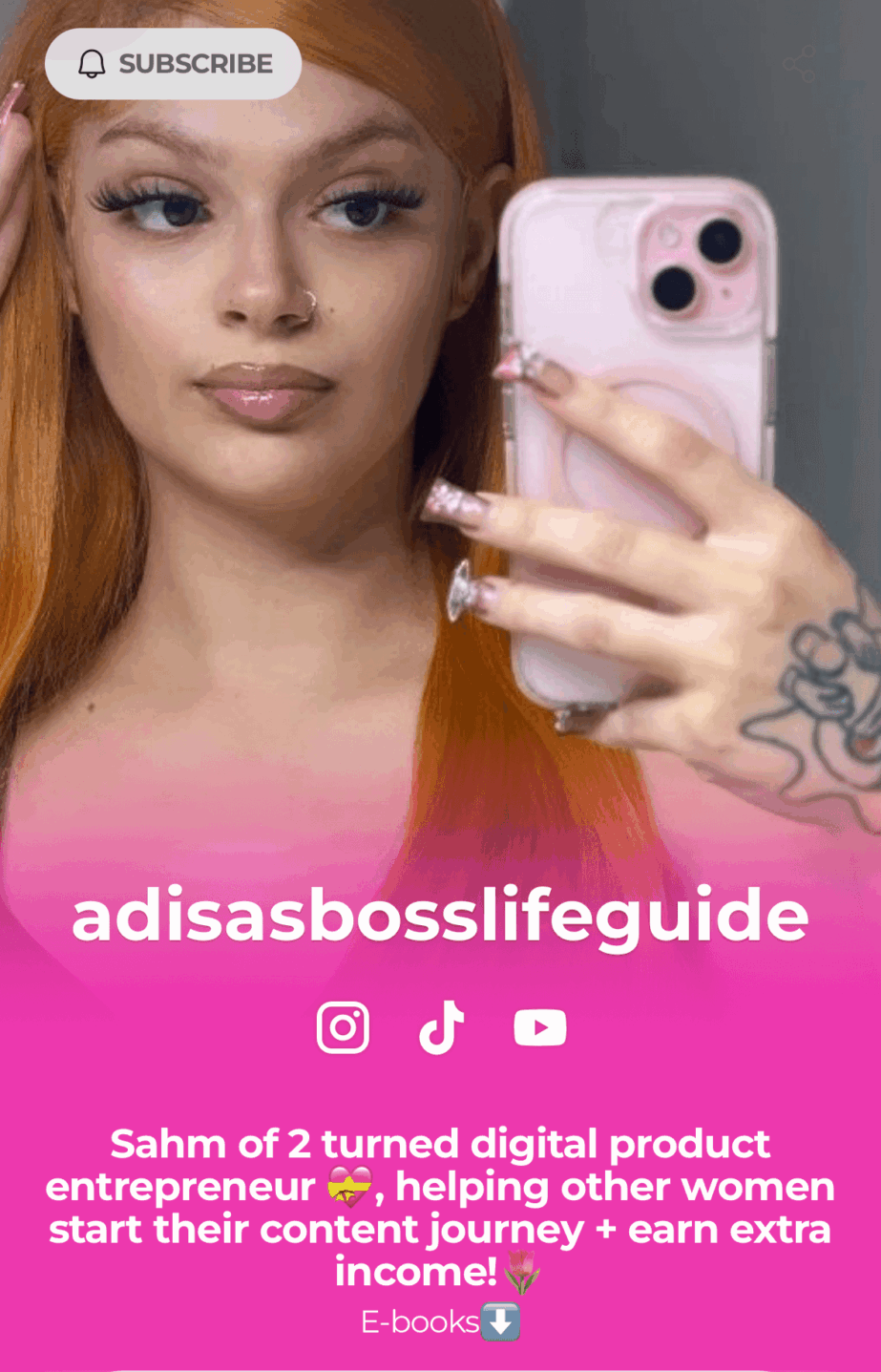 Young woman with long red hair taking a mirror selfie with a pink phone, wearing a pink top, with a tattoo on her hand.