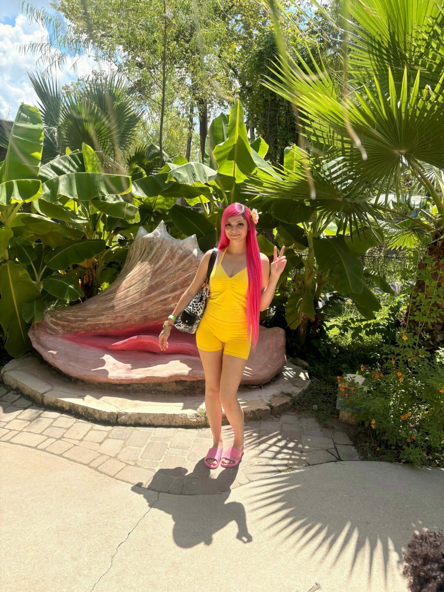Person with pink hair in yellow outfit standing on a stone path next to a large red and white shell sculpture, surrounded by greenery.