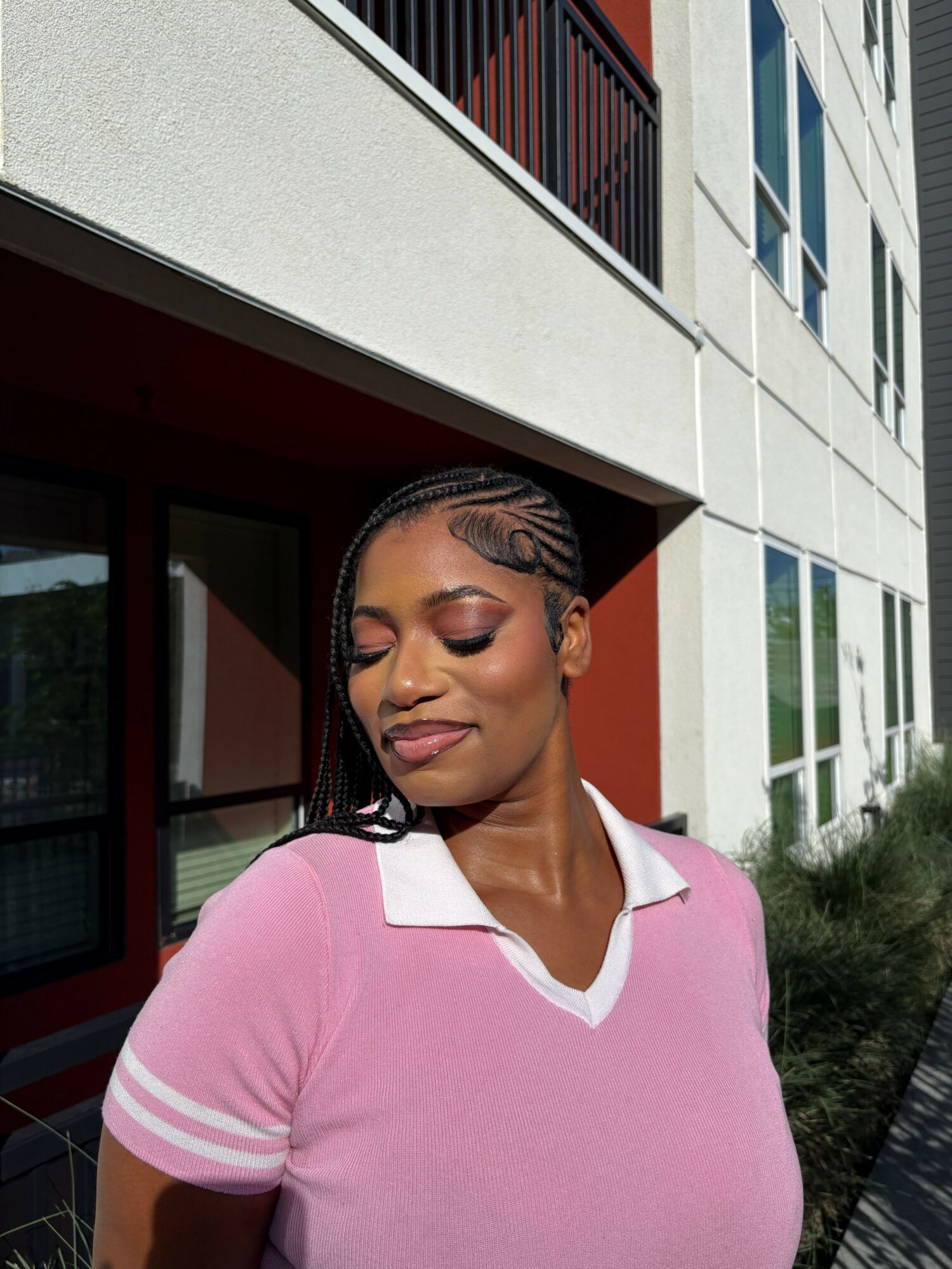 Young woman with braided hair and closed eyes, wearing a pink and white shirt, standing outside a building.