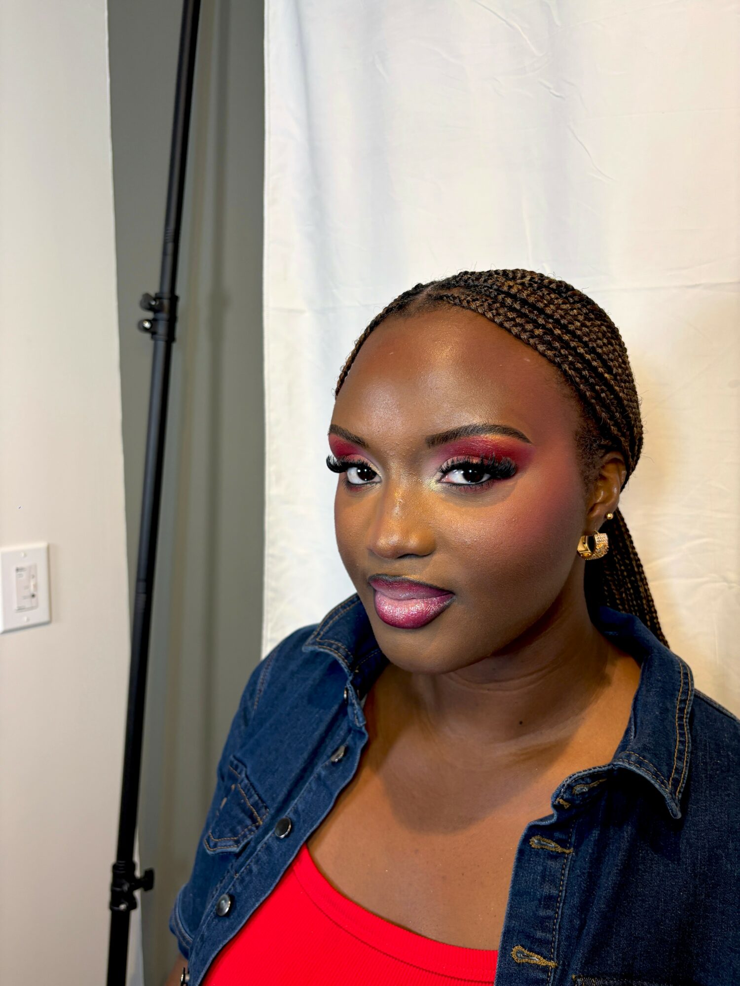 Portrait of a woman with braided hair, wearing a red top and denim jacket, standing in front of a white backdrop.