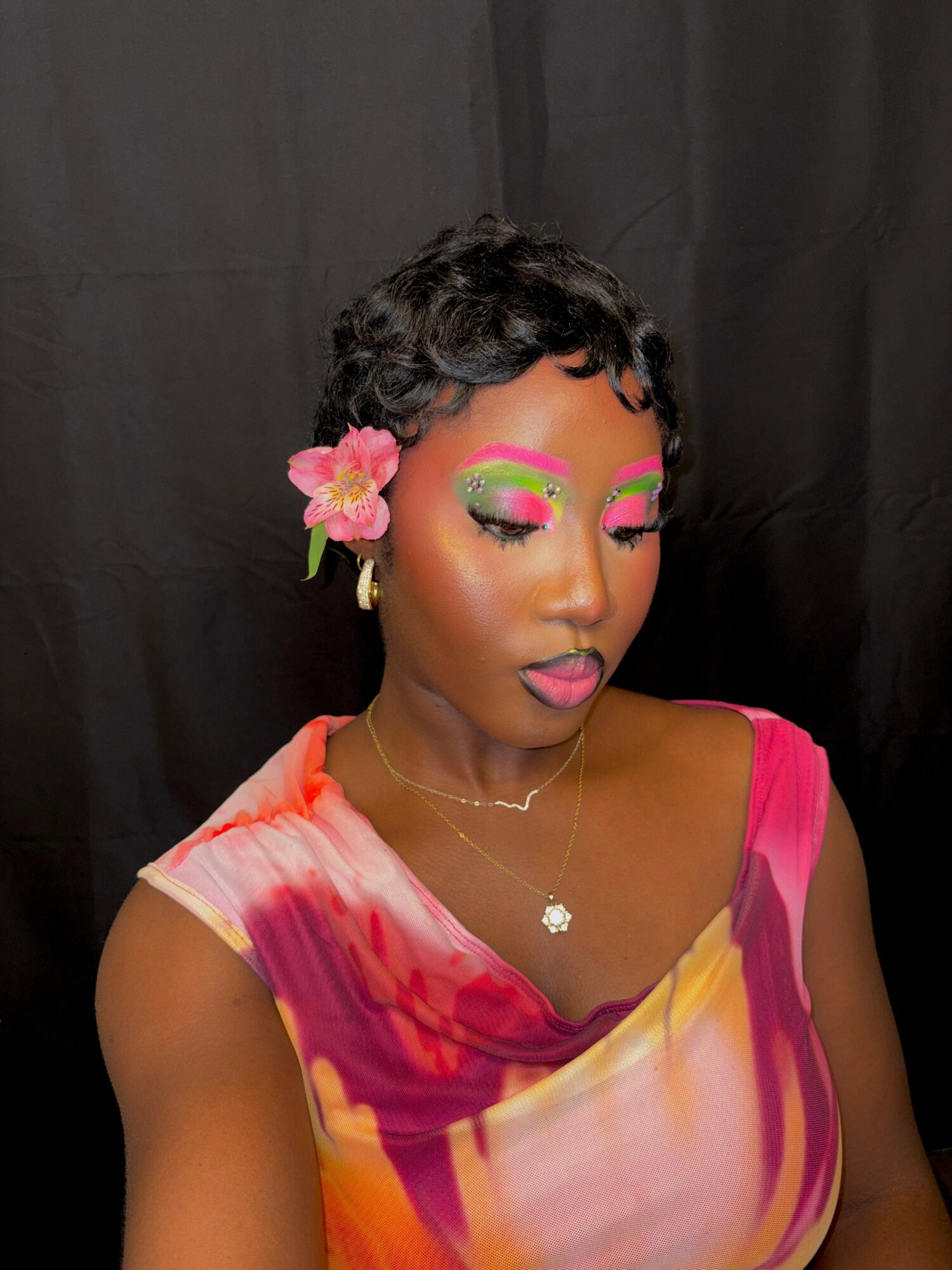Woman with short curly hair, pink flower in hair, colorful makeup, wearing a vibrant pink and yellow dress, against black background.