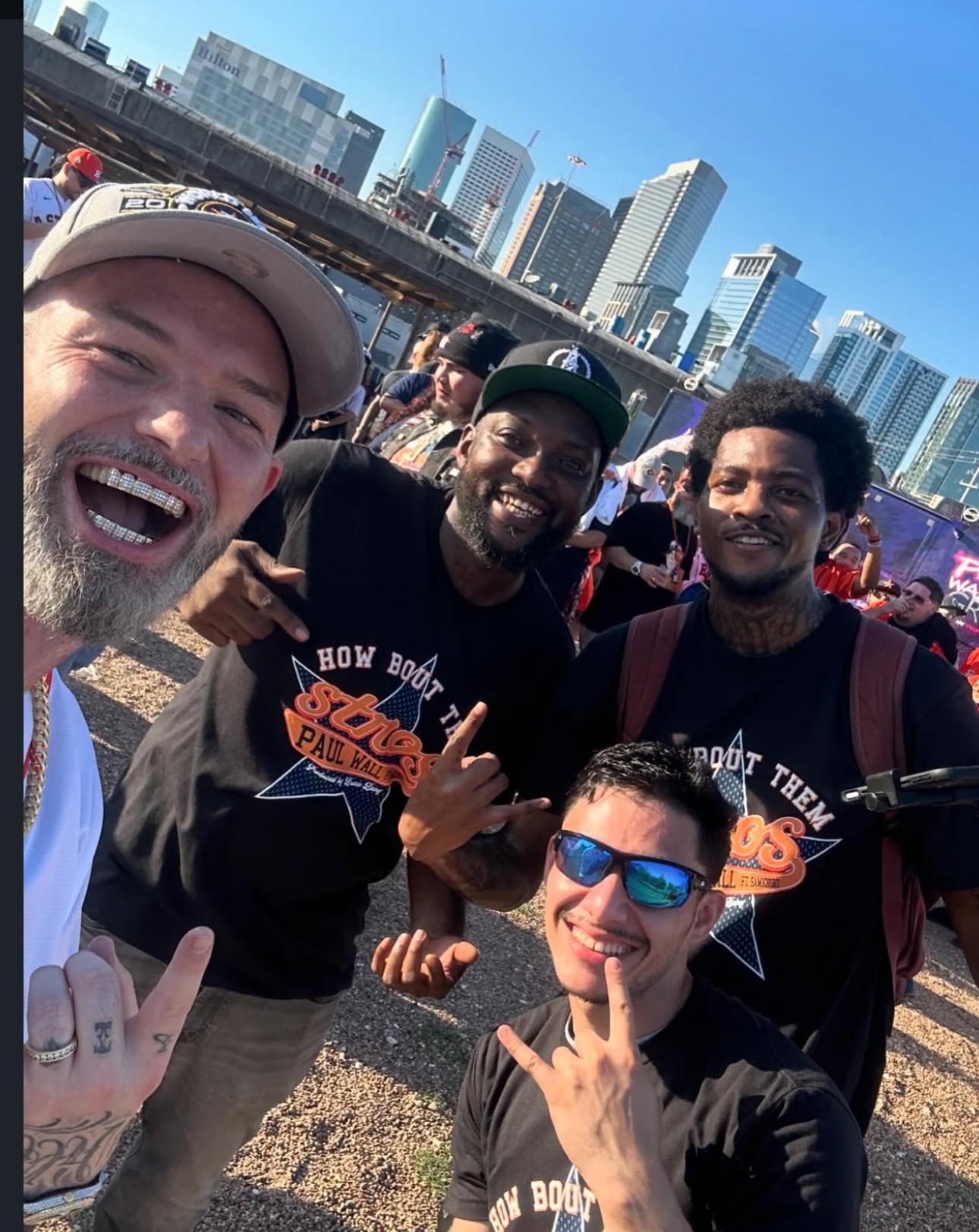 Four people smiling outdoors with city skyline in background, one wearing sunglasses, two making hand gestures, sunny day.