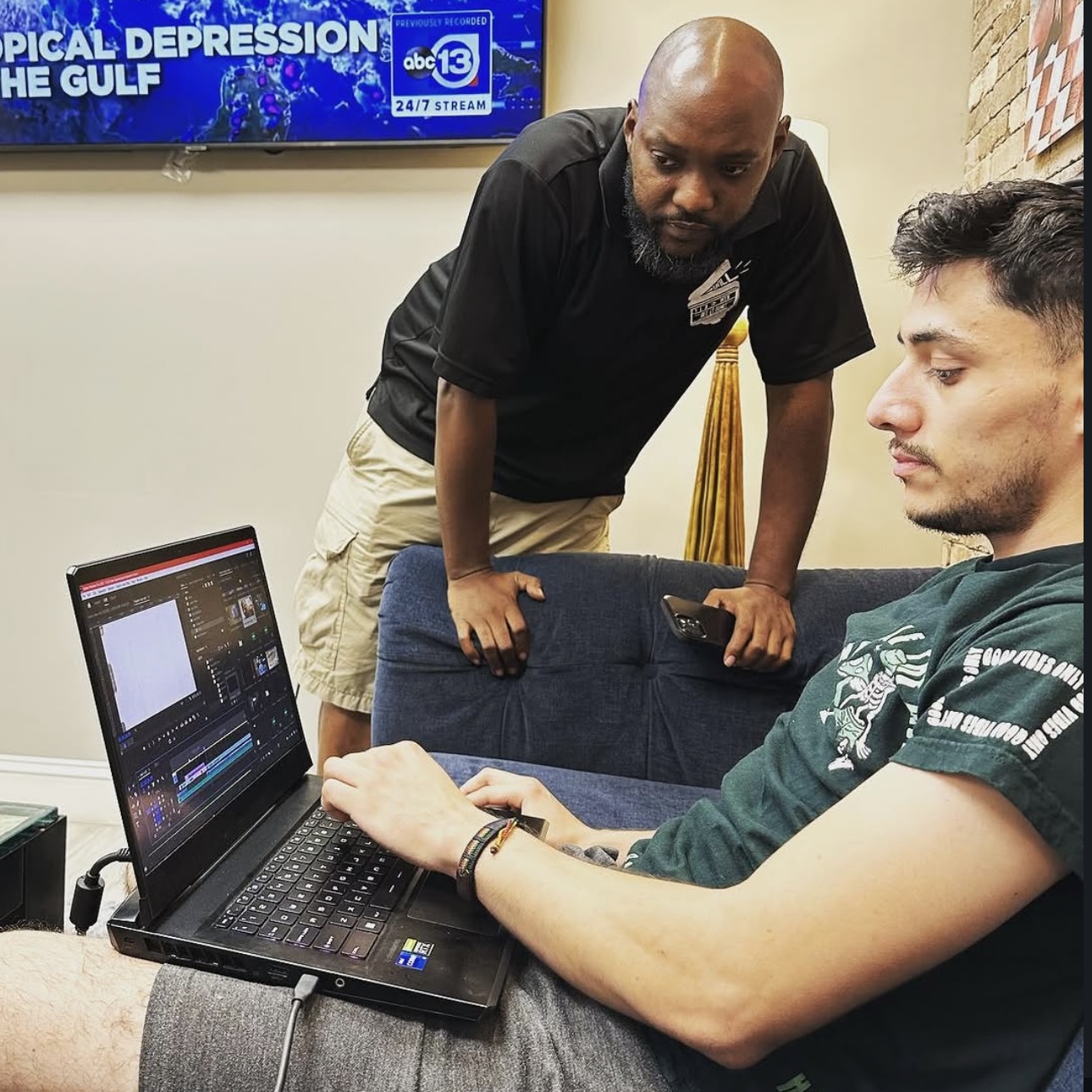 Two men in a room, one sitting with a laptop and the other standing, looking at the screen. A TV displays news in the background.