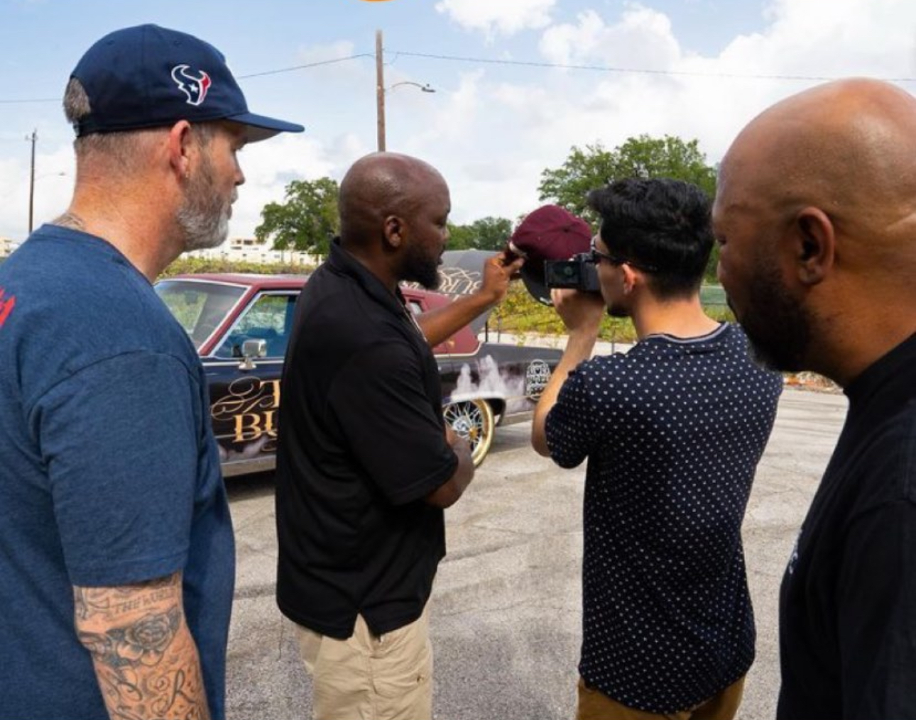 Four people standing outdoors, one holding a camera, with a car and trees in the background.
