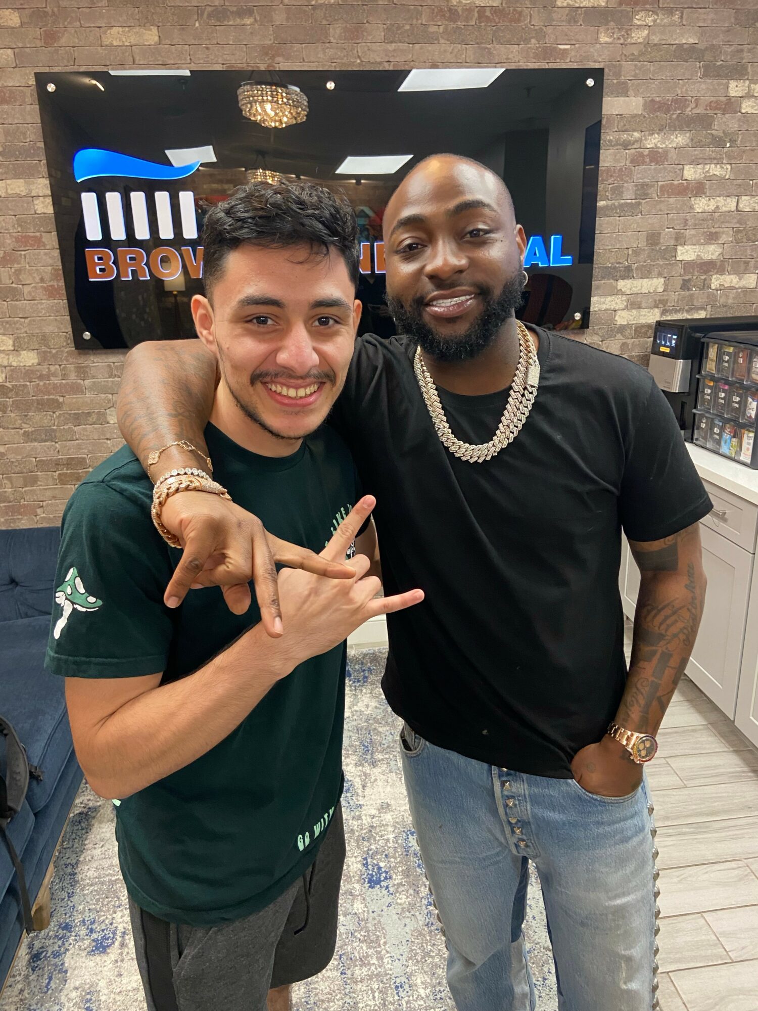 Two men smiling and posing together indoors, one making a hand gesture, with a brick wall and a TV screen in the background.